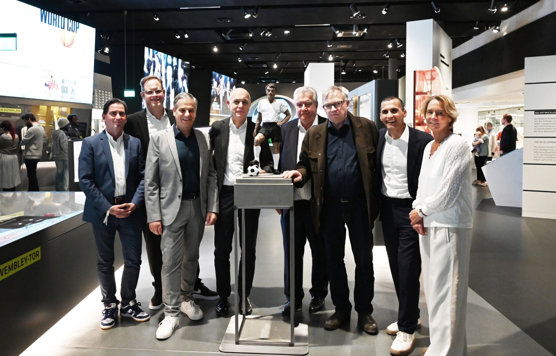 [{"clang_id":1,"value":"Gruppe von acht erwachsenen Personen steht lächelnd in einem Museum vor einer Ausstellung über Fußball. Im Hintergrund weitere Museumsbesucher und Exponate."}]