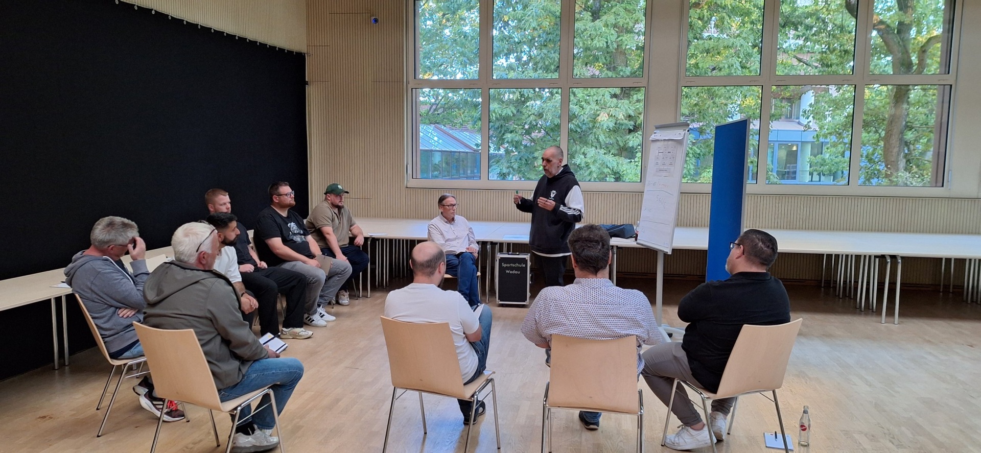 Versammlung in einem hellen Raum. Neun Männer sitzen im Kreis auf Stühlen, ein Mann steht und spricht, Papier an der Wand.