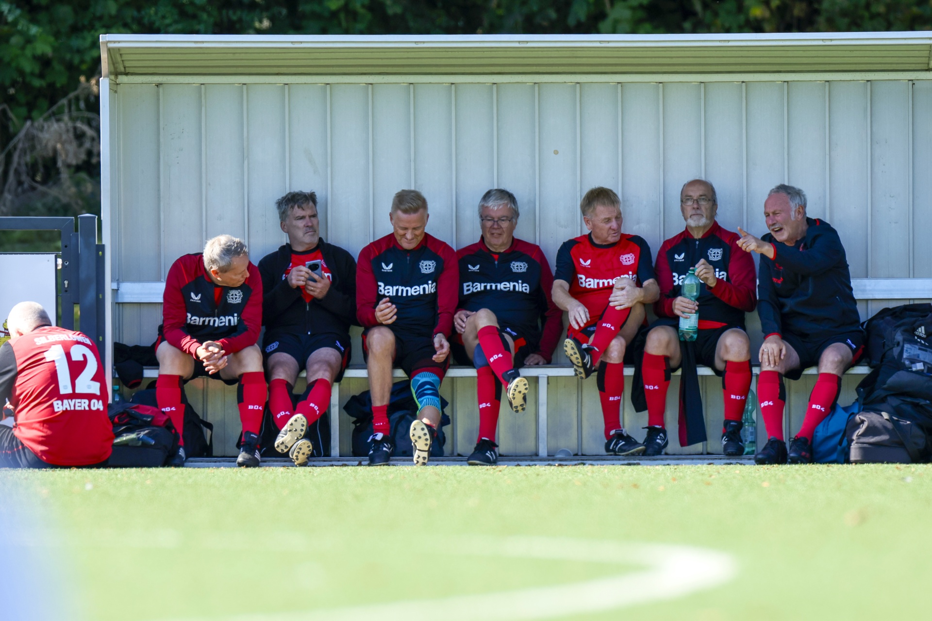 [{"clang_id":1,"value":"Fußballmannschaft auf einer Bank: Sieben Männer in roten und schwarzen Trainingsanzügen sitzen auf einer weißen Bank am Spielfeldrand. Im Hintergrund grüne Bäume."}]
