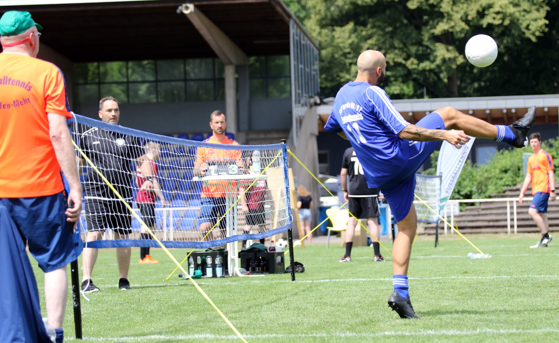 [{"clang_id":1,"value":"Fußvolleyballspiel: Spieler in blau schlägt Ball über Netz. Im Hintergrund Zuschauer und Gebäude, grüne Rasenfläche."}]
