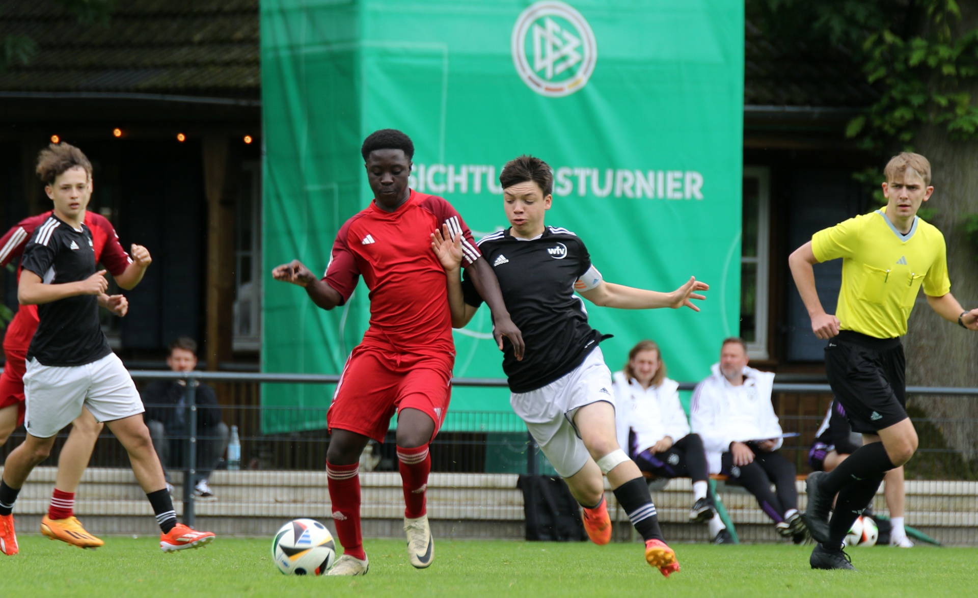 [{"clang_id":1,"value":"Fußballspiel: Drei Jugendspieler kämpfen um den Ball. Links ein Spieler in Schwarz-Weiß, Mitte in Rot, rechts in Gelb. Im Hintergrund Banner und Zuschauer."}]