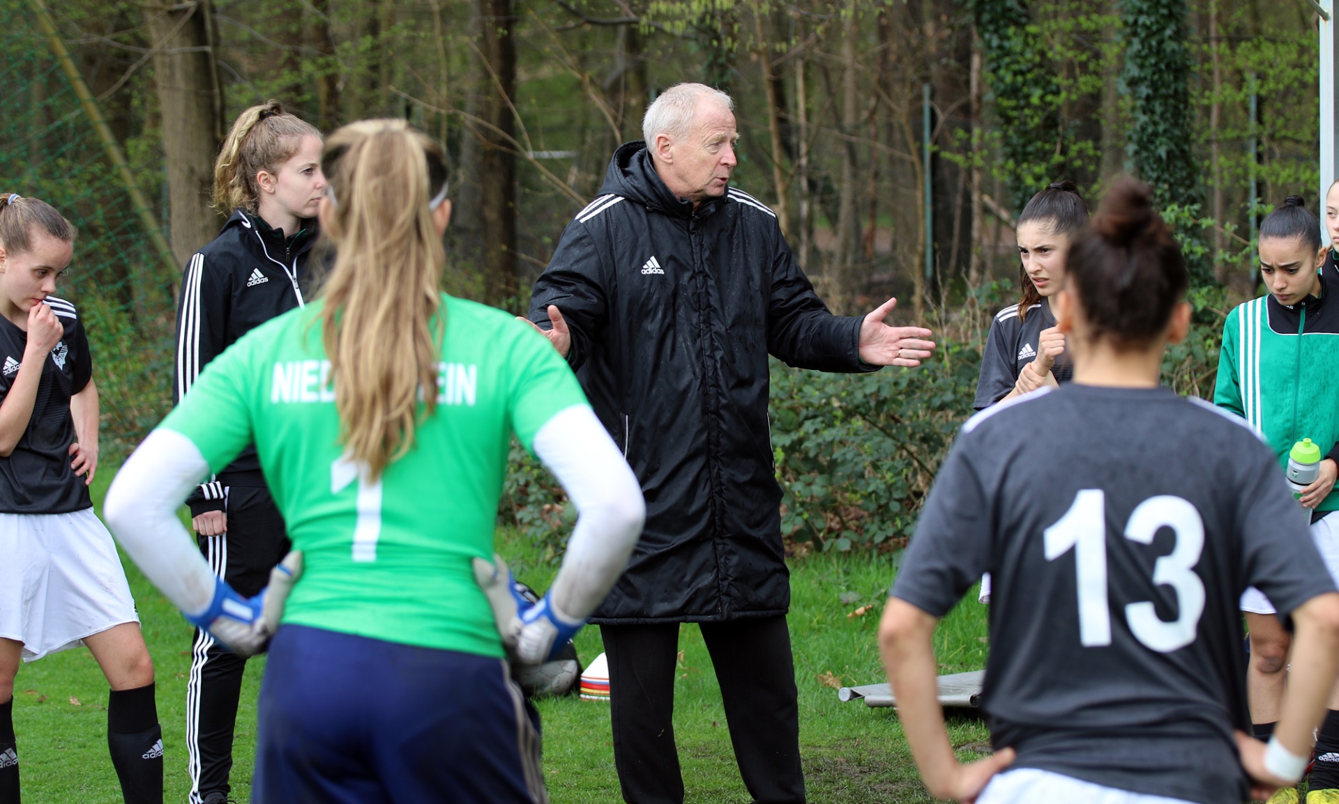 [{"clang_id":1,"value":"Fußballtrainer steht vor einer Gruppe von Spielerinnen im Trainingsoutfit. Er gestikuliert, die Spielerinnen schauen ihn an. Im Hintergrund Bäume."}]