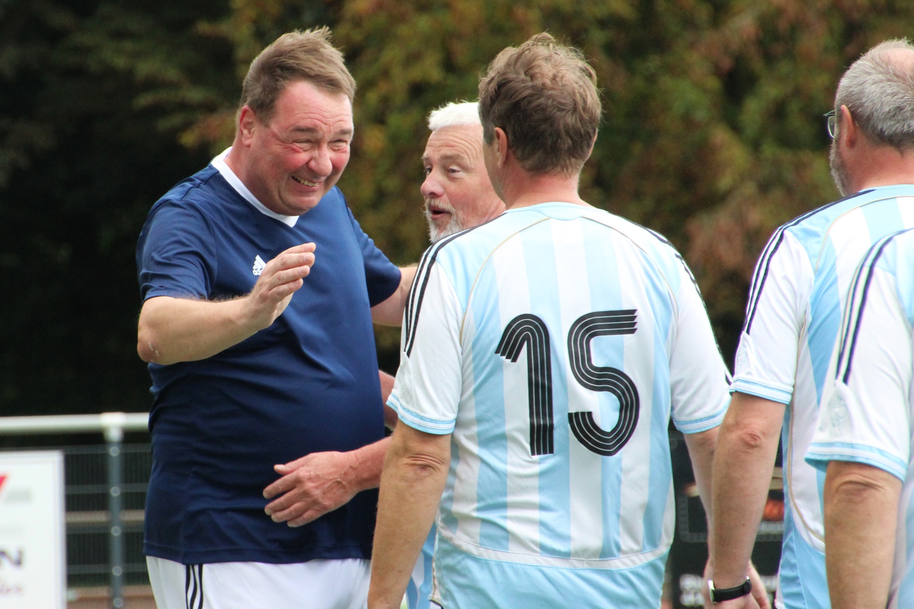 [{"clang_id":1,"value":"Drei ältere Männer auf einem Fußballfeld. Links ein Mann in blauem Trikot, der mit einem Mann in blau-weiß gestreiftem Trikot mit Rückennummer 15 spricht. Im Hintergrund ein weiterer Spieler."}]