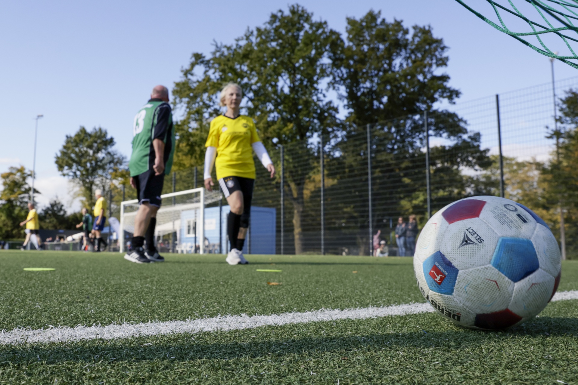 [{"clang_id":1,"value":"Fußballfeld mit Spielern im Hintergrund. Im Vordergrund ein Fußball auf dem grünen Rasen. Netz und Bäume im Hintergrund."}]