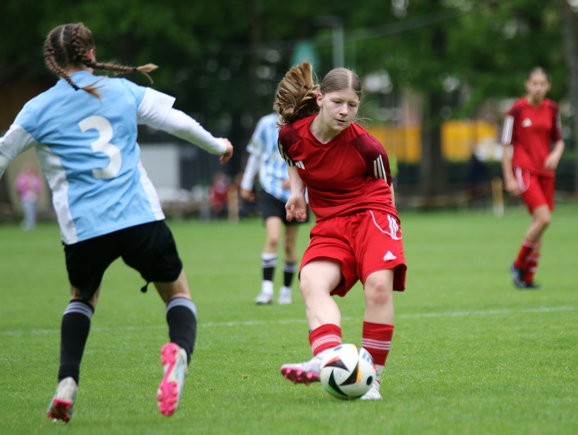 [{"clang_id":1,"value":"Fußballspiel: Mädchen in roten Trikots dribbelt den Ball auf grünem Rasen, verfolgt von Gegenspielerin mit blauem Trikot. Hintergrund verschwommen."}]