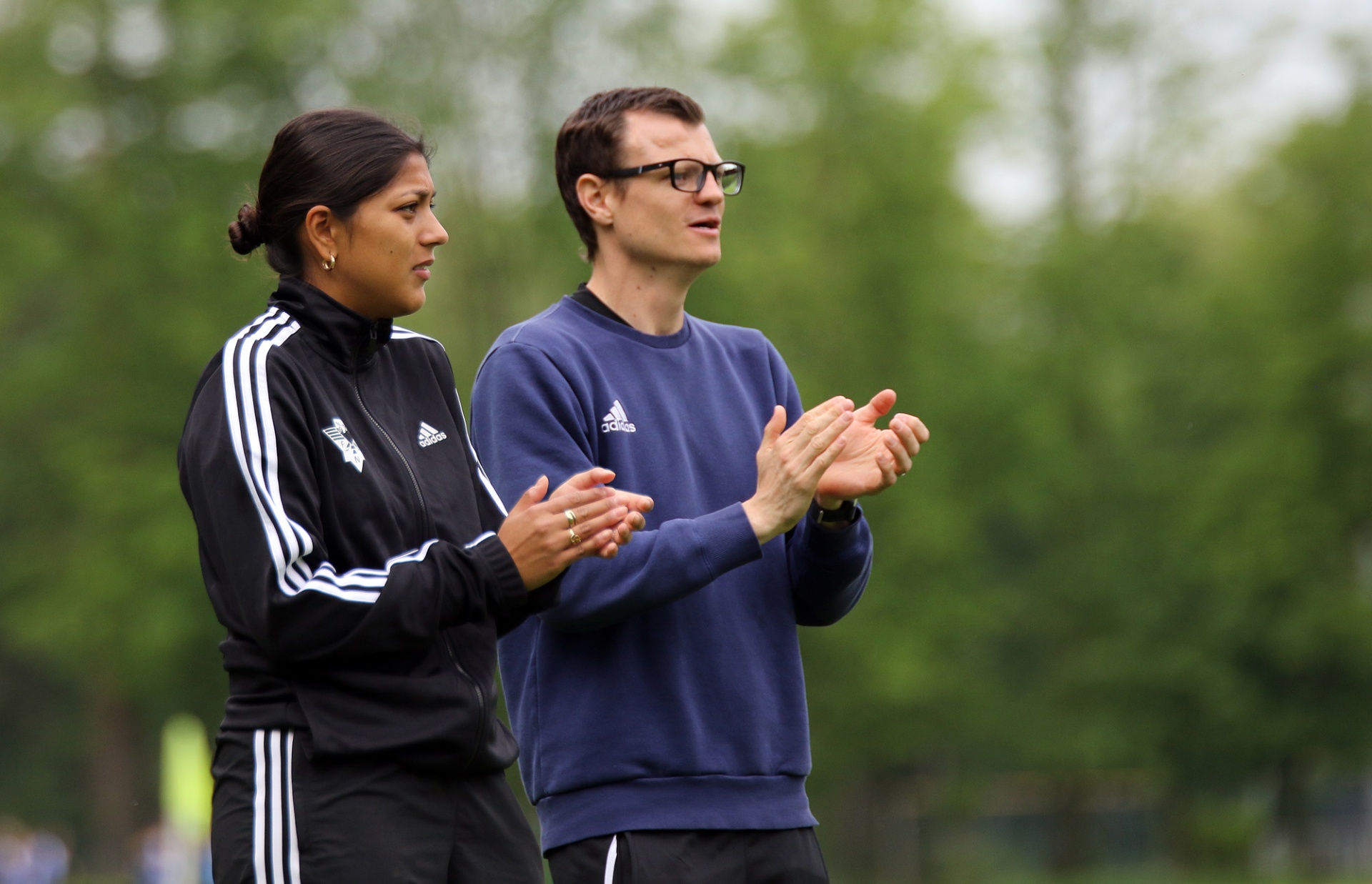 [{"clang_id":1,"value":"Zwei Personen, eine Frau mit dunklen Haaren und ein Mann mit Brille, klatschen im Freien vor grünem Hintergrund. Beide tragen Sportkleidung mit Adidas-Logo."}]