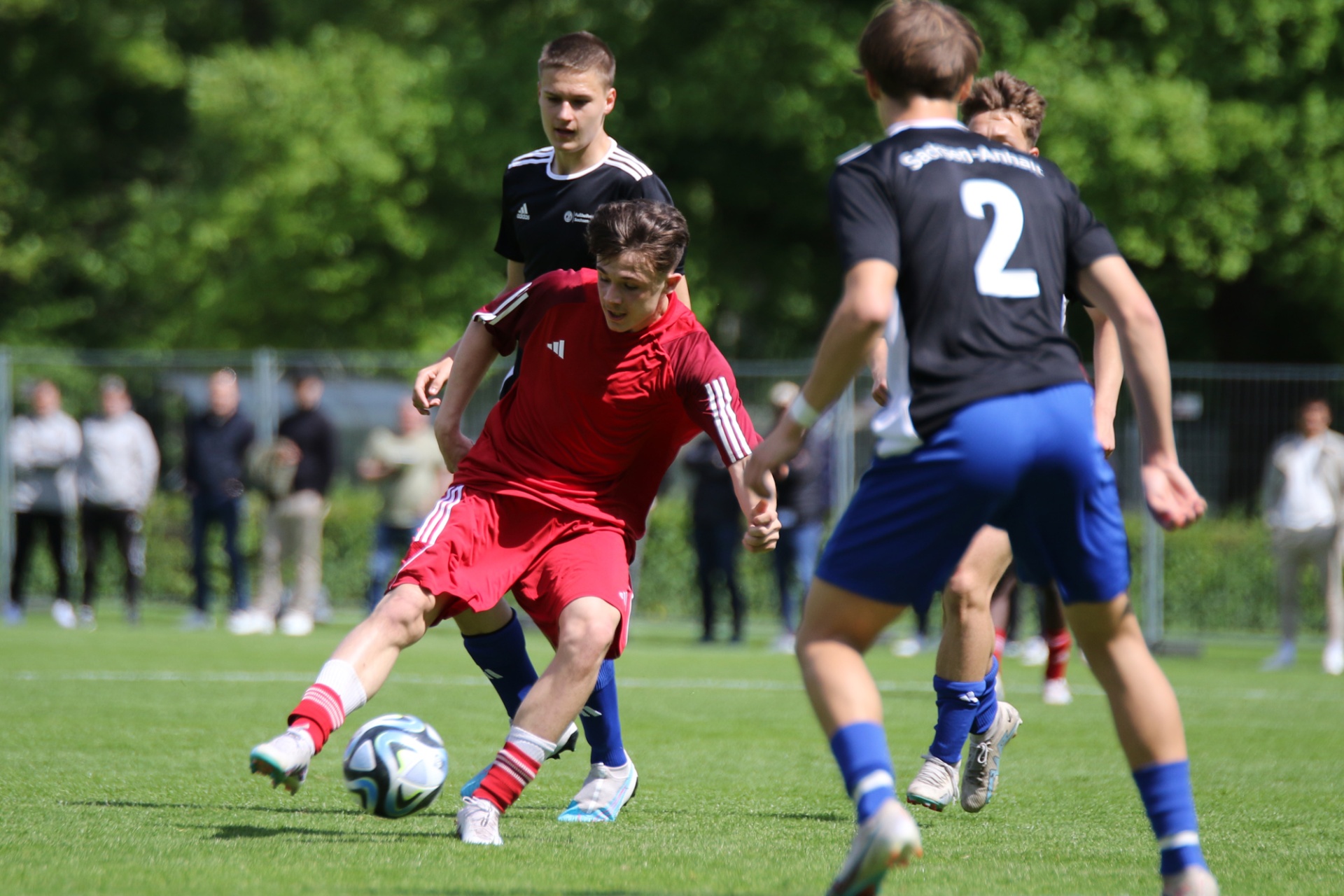 [{"clang_id":1,"value":"Fußballspiel: Jugendlicher in Rot dribbelt den Ball, verfolgt von zwei Gegenspielern in Blau und Schwarz. Im Hintergrund Zuschauer und Bäume."}]