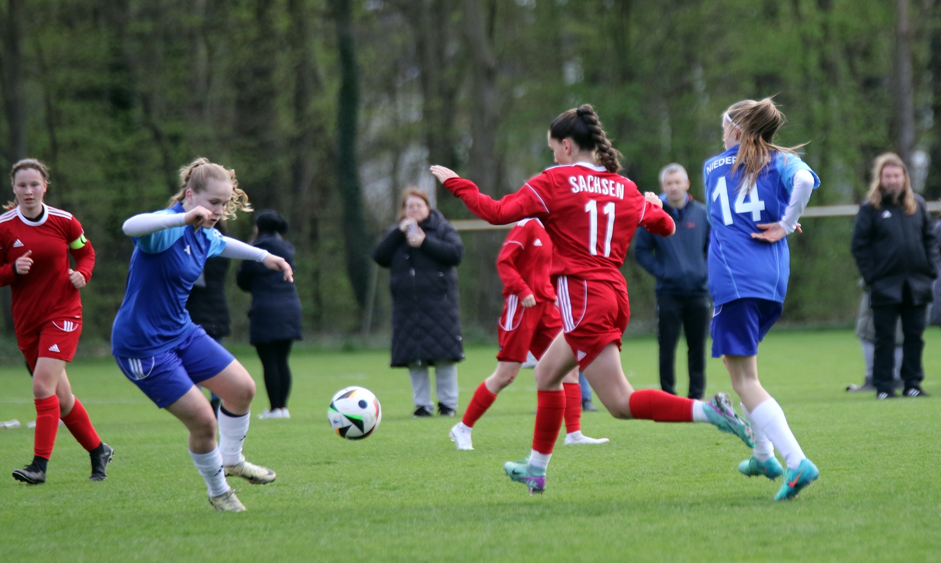 [{"clang_id":1,"value":"Fußballspiel: Spielerinnen in Rot und Blau kämpfen um den Ball auf einem grünen Rasen. Zuschauer im Hintergrund."}]