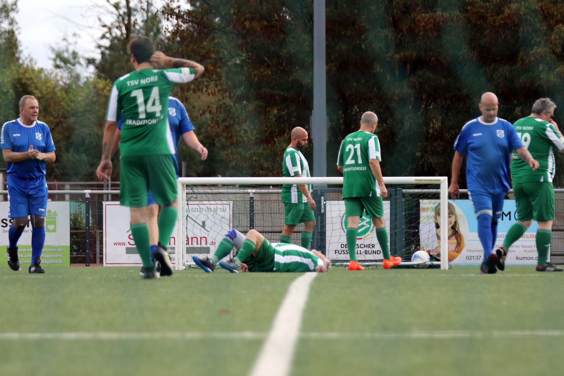 [{"clang_id":1,"value":"Fußballspiel: Spieler in Grün und Blau, einer springt über am Boden liegenden Gegner. Im Hintergrund Zuschauer und Werbebanner. Rasenplatz."}]