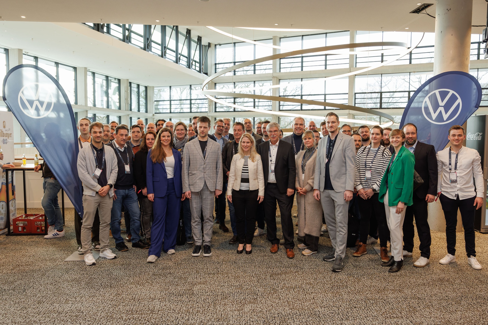 [{"clang_id":1,"value":"Gruppe von etwa 30 beruflich gekleideten Personen vor Volkswagen-Logos in einem hellen, modernen Raum mit Glasfassade."}]