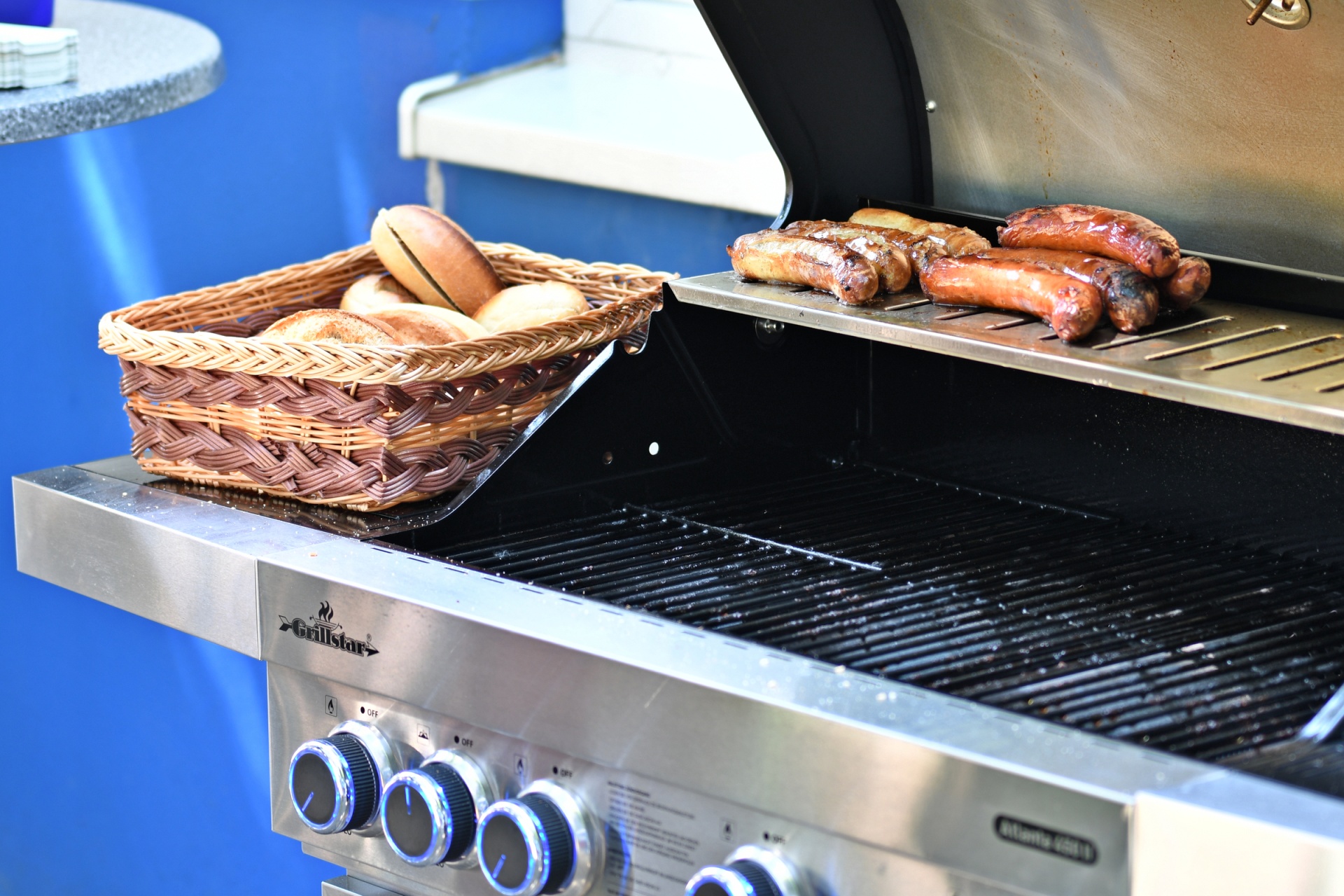 [{"clang_id":1,"value":"Grill mit Bratwürsten und einem Korb Brötchen darauf. Edelstahlgrill mit vier Reglern und Grillrost. Blauer Hintergrund."}]