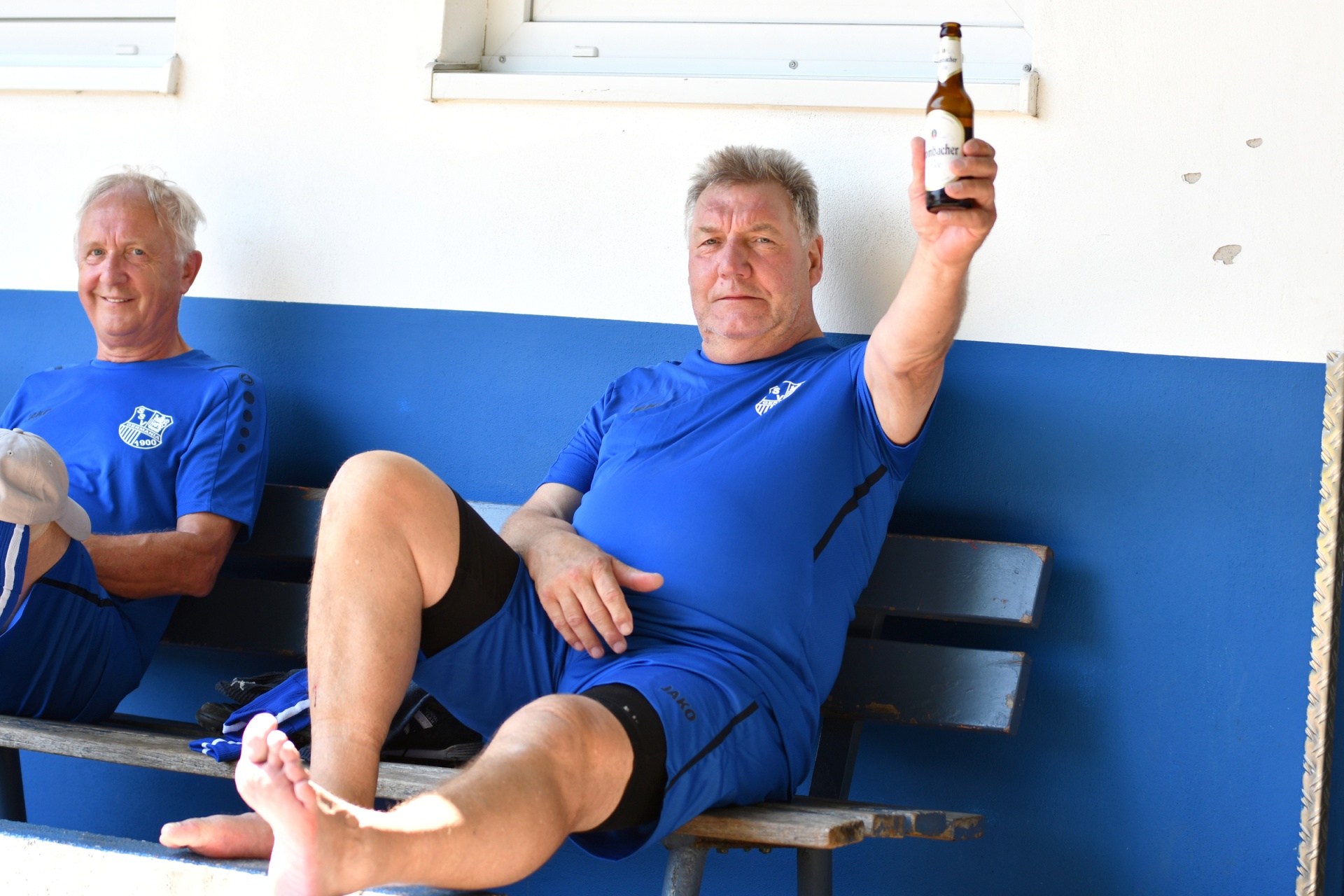 [{"clang_id":1,"value":"Zwei ältere Männer sitzen auf einer blauen Bank vor einer weißen Wand. Einer hält eine Bierflasche hoch, beide tragen blaue Sportkleidung."}]