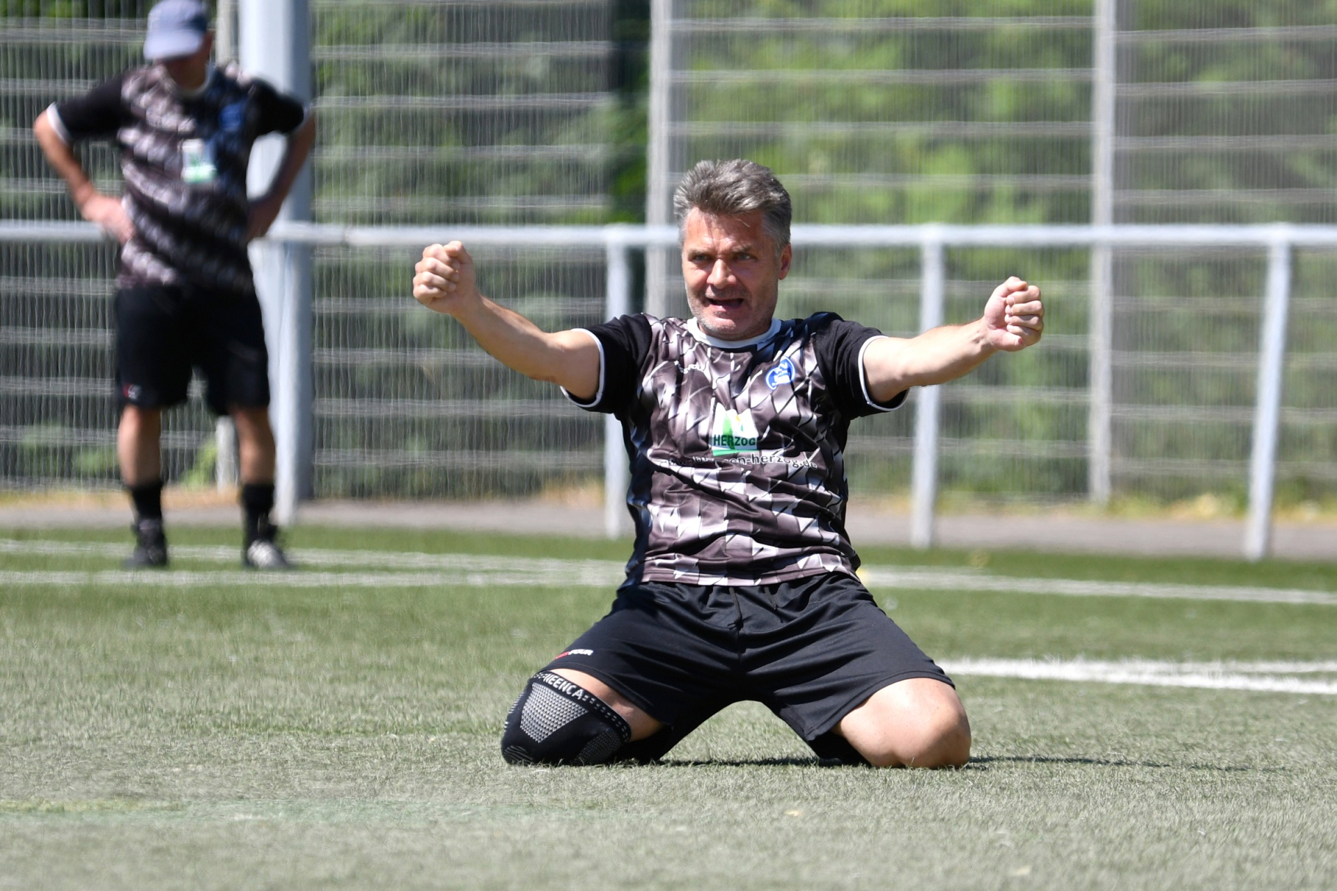 [{"clang_id":1,"value":"Mann kniet auf grünem Rasen, Arme weit geöffnet, jubelnd. Trägt Fußballtrikot und -shorts. Im Hintergrund ein Zaun und verschwommene Personen."}]