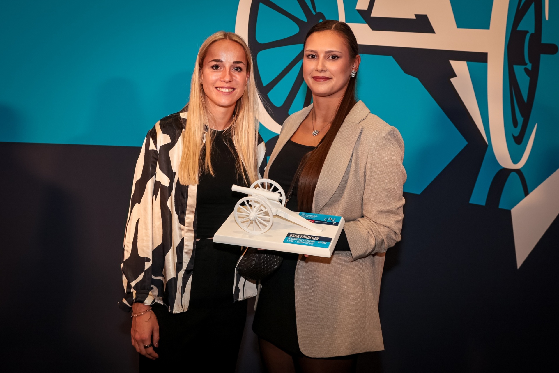 [{"clang_id":1,"value":"Zwei Frauen posieren lächelnd mit einer Auszeichnung in Form eines Fahrrads. Eine trägt eine Jacke mit Muster, die andere eine graue Jacke. Dunkler Hintergrund mit blauem Grafikmotiv."}]