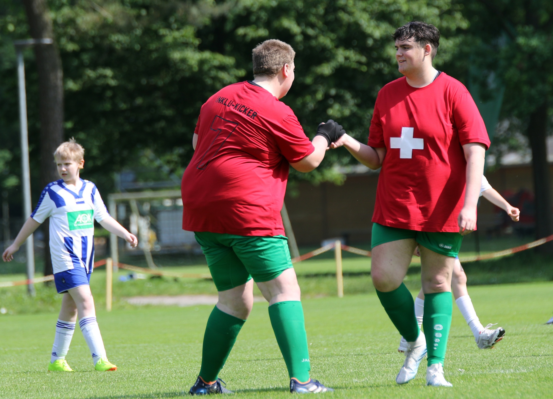 [{"clang_id":1,"value":"Zwei Fußballspieler in rot-grün, einer mit Aufschrift \"Mr. Lu-Kicker\", geben sich die Hände. Im Hintergrund ein Kind und ein Fußballfeld."}]