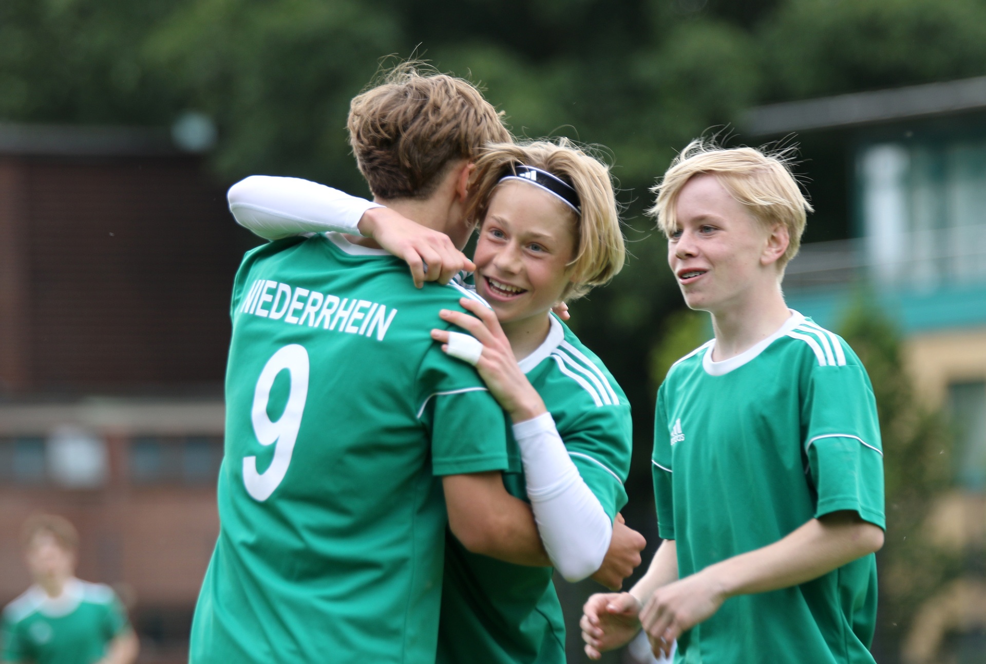 [{"clang_id":1,"value":"Drei junge Fußballspieler in grünen Trikots umarmen sich freudig auf einem Fußballfeld. Im Hintergrund ein Gebäude und Grünflächen."}]