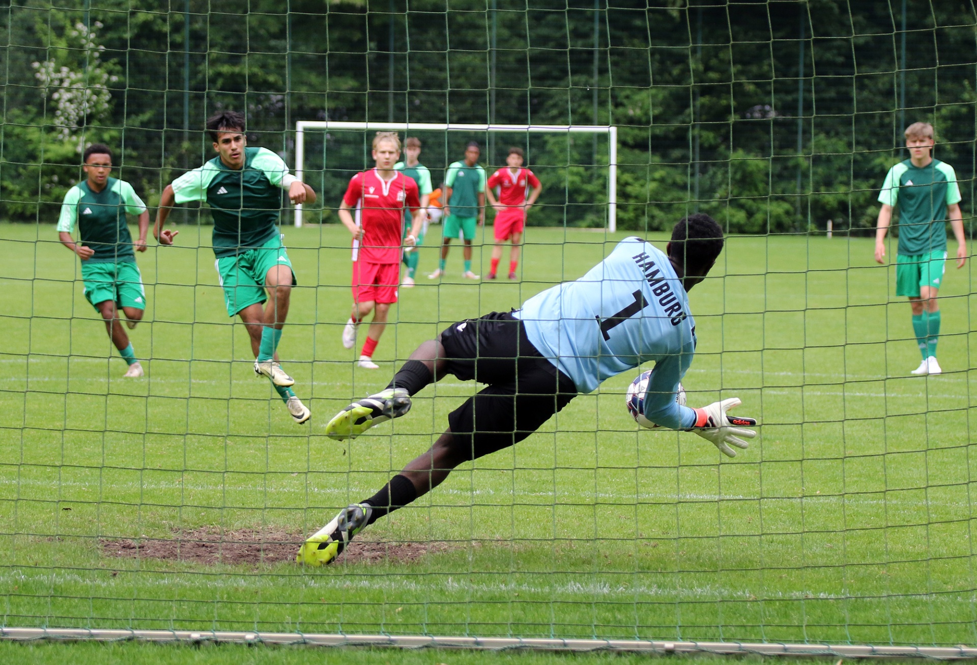 [{"clang_id":1,"value":"Fußballspiel: Torwart in schwarzer Kleidung springt nach rechts, um Ball zu halten. Zwei Spieler in grün und rot im Hintergrund. Rasenplatz und Netz im Vordergrund."}]