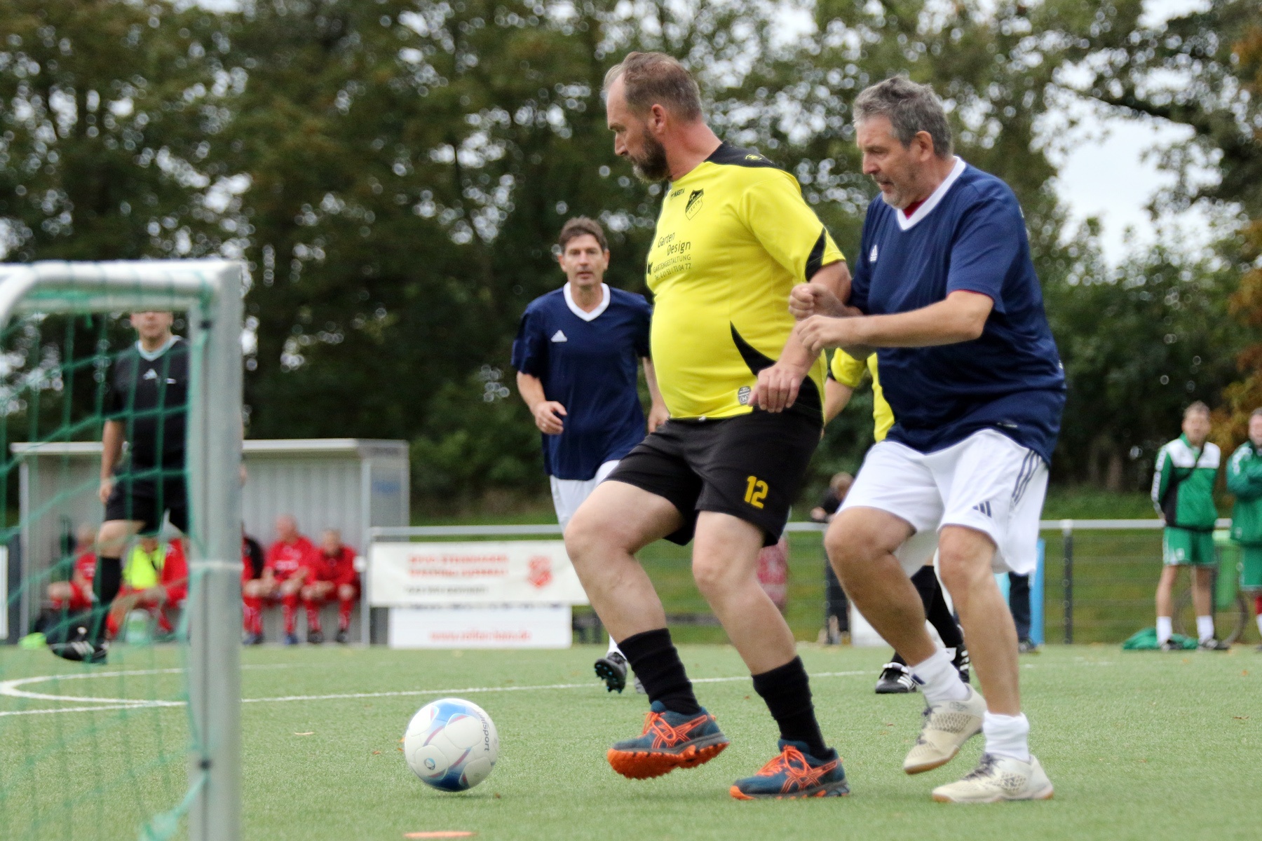 [{"clang_id":1,"value":"Zwei Männer spielen Fußball auf einem grünen Kunstrasenplatz. Im Hintergrund weitere Spieler und ein Fußballtor."}]