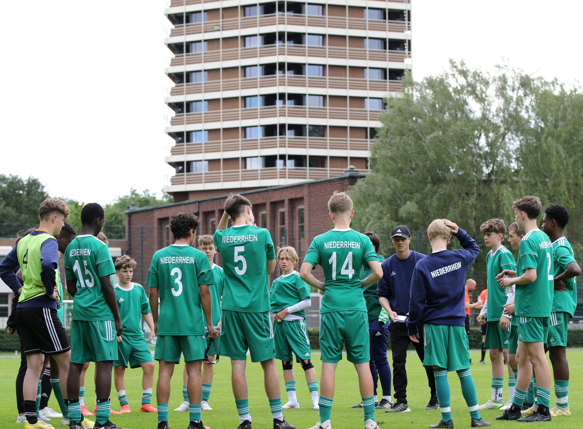 [{"clang_id":1,"value":"Fußballteam in grünen Trikots steht auf Rasen vor einem mehrstöckigen Gebäude. Ein Trainer erklärt etwas, Spieler hören zu."}]