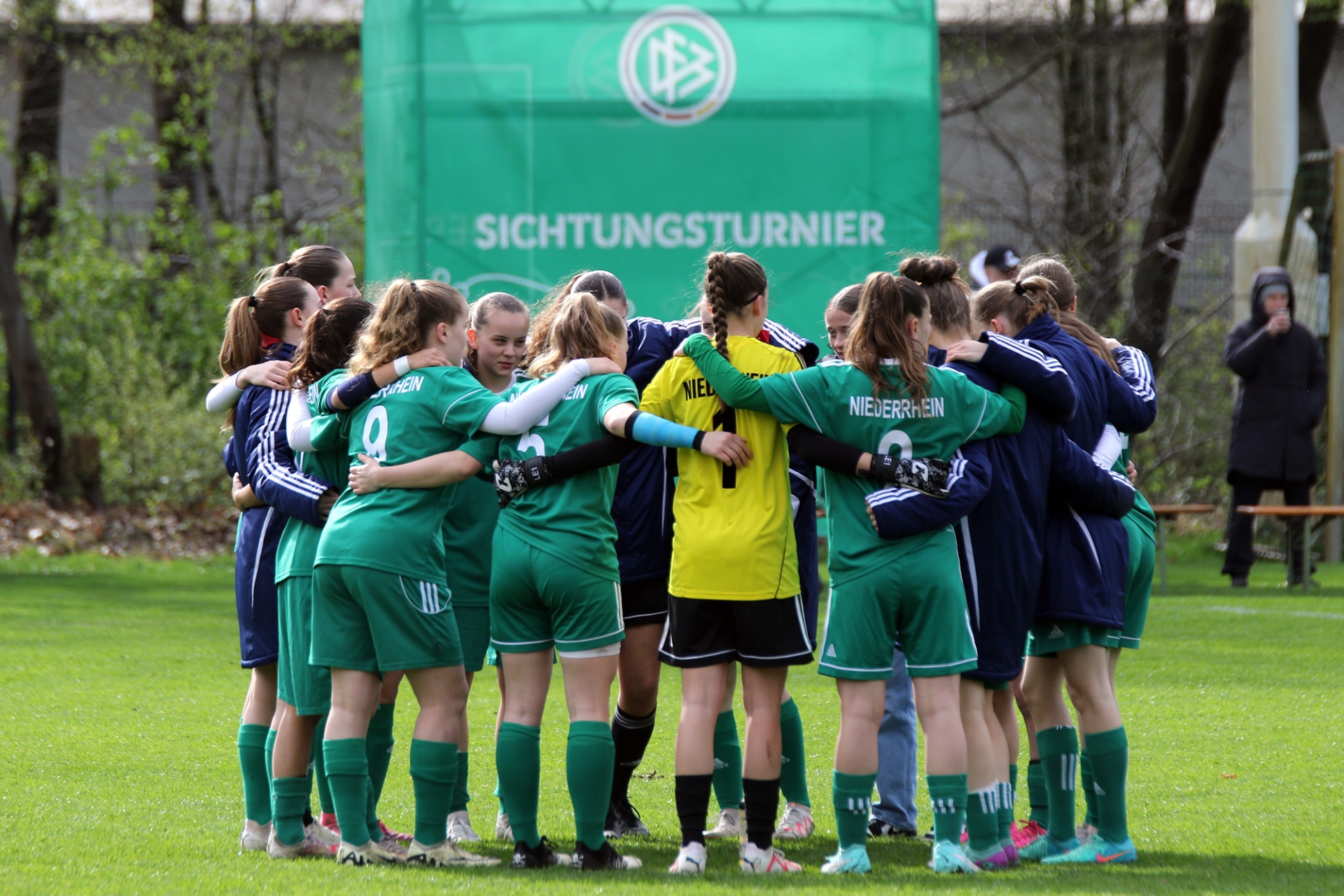[{"clang_id":1,"value":"Fußballmannschaft im Kreis huddle auf Rasenfeld. Spielerinnen tragen grüne und blaue Trikots mit Vereinslogo. Im Hintergrund Banner mit Schriftzug \"SICHTUNGSTURNIER\"."}]