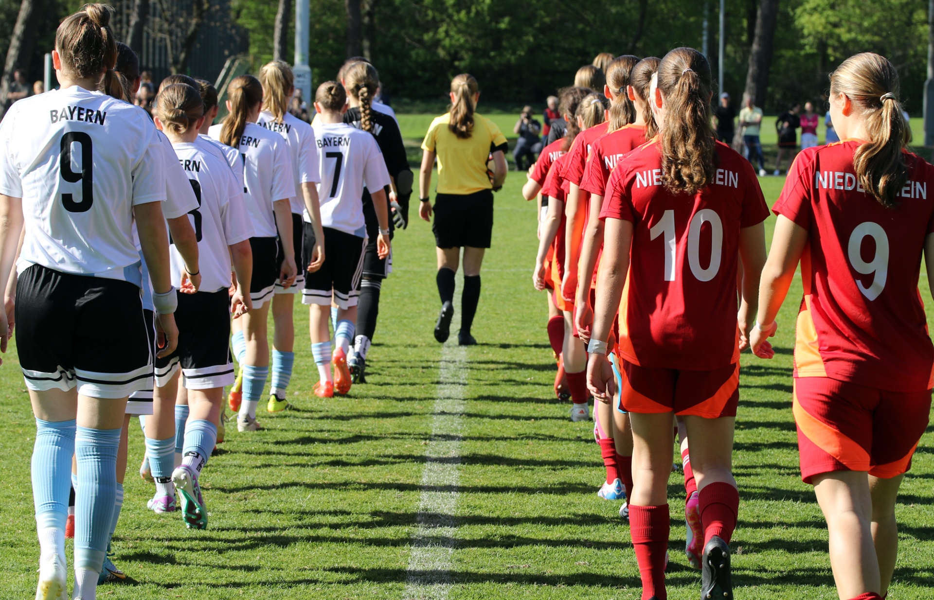 [{"clang_id":1,"value":"Zwei Fußballmannschaften in weißen und roten Trikots stehen sich auf einem grünen Rasenfeld gegenüber. Ein Schiedsrichter steht zwischen ihnen."}]