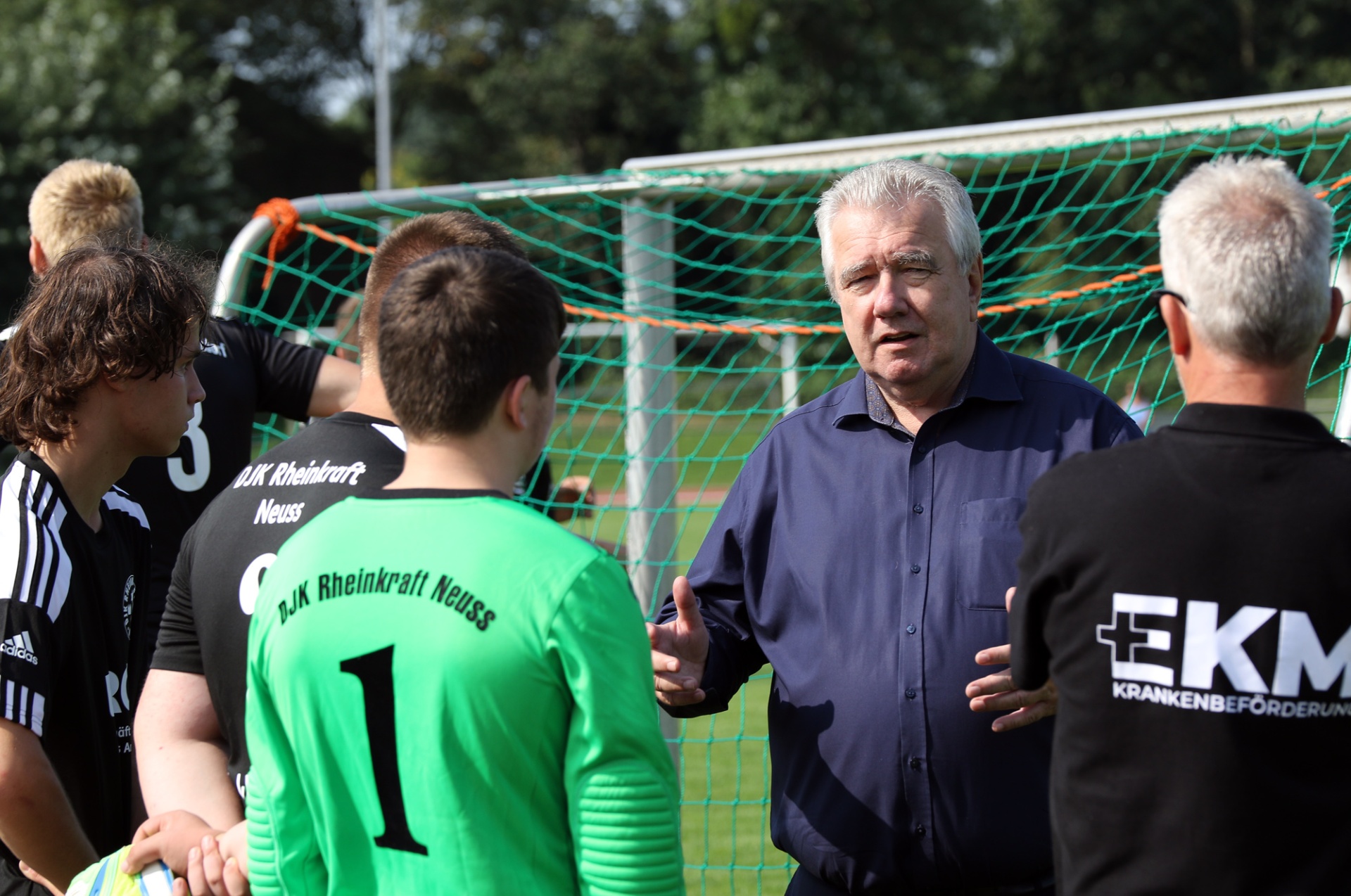 [{"clang_id":1,"value":"Fußballtrainer mit grauem Haar und blauem Hemd spricht mit drei Spielern vor einem Fußballtor. Im Hintergrund ein Mann mit schwarzer Weste und Logo \"EKM\"."}]