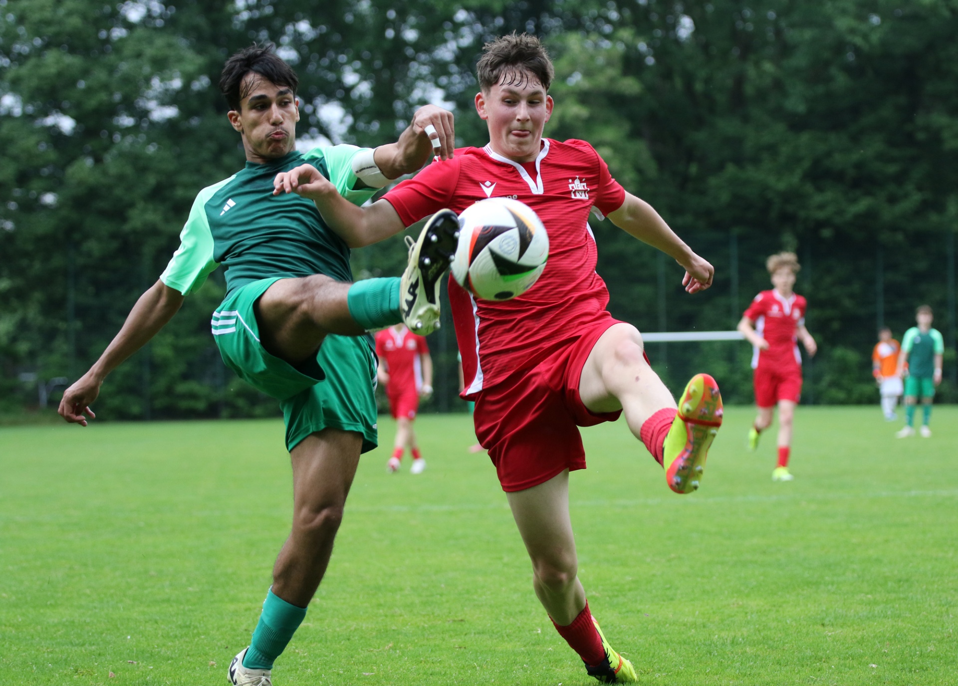 [{"clang_id":1,"value":"Zwei Jugendliche spielen Fußball auf einem Rasenplatz. Links ein Spieler in grünem Trikot, rechts ein Spieler in rotem Trikot, der den Ball tritt. Im Hintergrund weitere Spieler und Bäume."}]