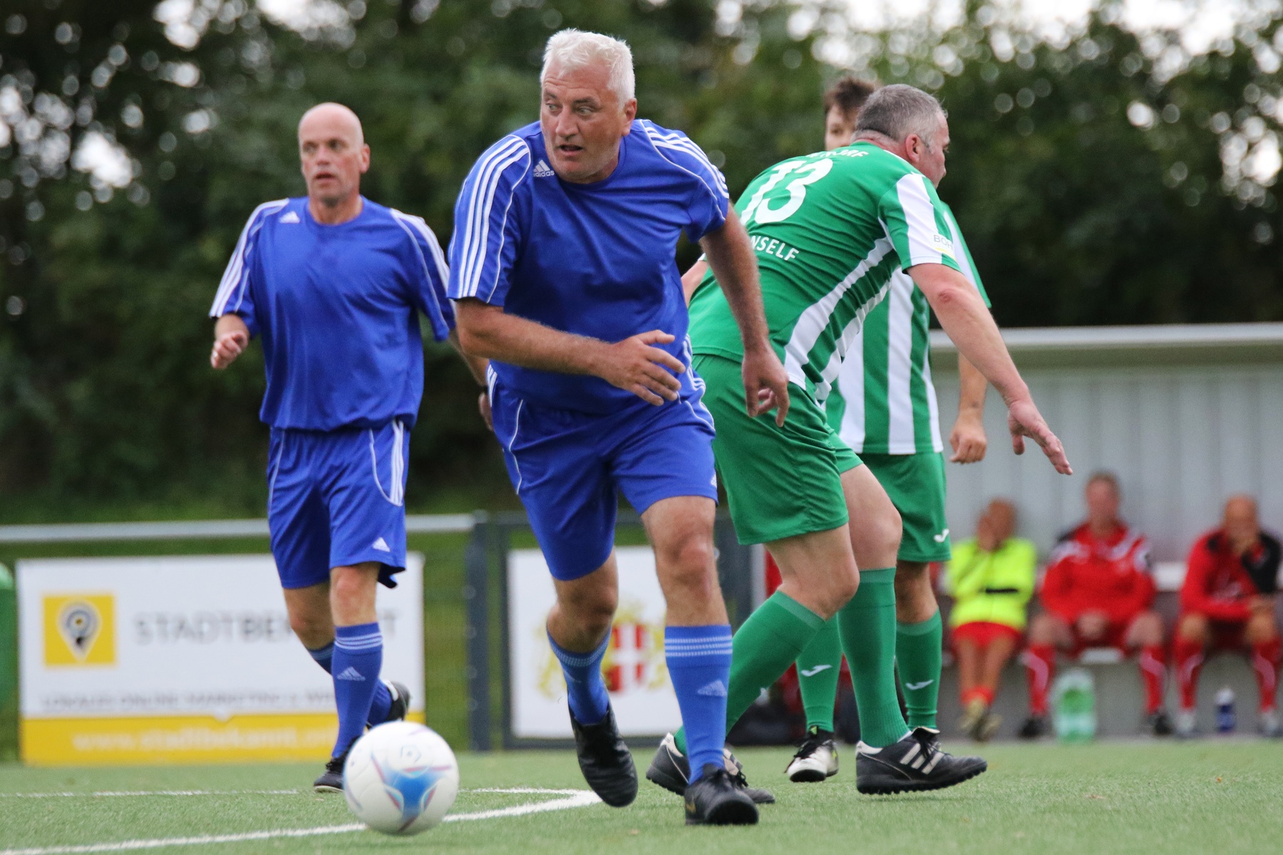 [{"clang_id":1,"value":"Fußballspiel: Zwei Männer in blau und ein Mann in grün-weiß kämpfen um den Ball auf einem Rasenplatz. Zuschauer im Hintergrund."}]