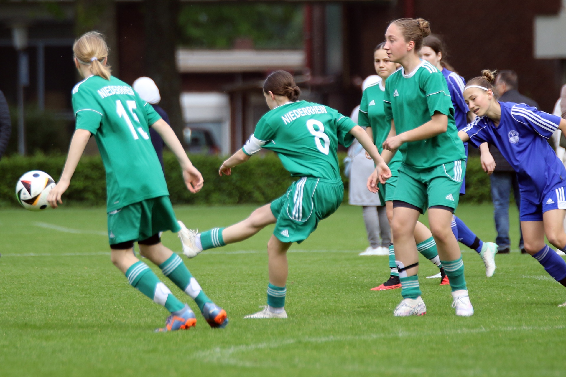 [{"clang_id":1,"value":"Fußballspiel mit jungen Spielerinnen. Spielerinnen in grünen und blauen Trikots auf Rasenfeld, eine schießt Ball. Gebäude im Hintergrund."}]