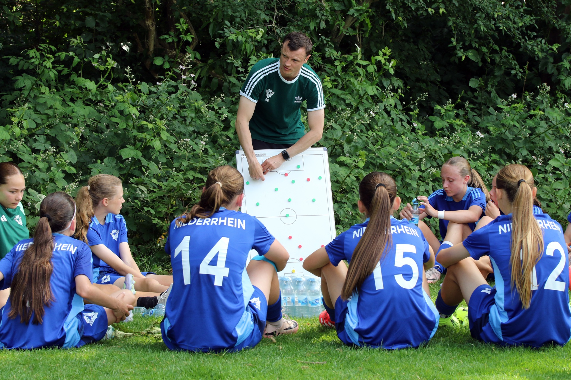 [{"clang_id":1,"value":"Fußballteam in blauem Trikot sitzt im Kreis auf Gras. Trainer erklärt Taktik an Whiteboard. Im Hintergrund Bäume und Sträucher."}]