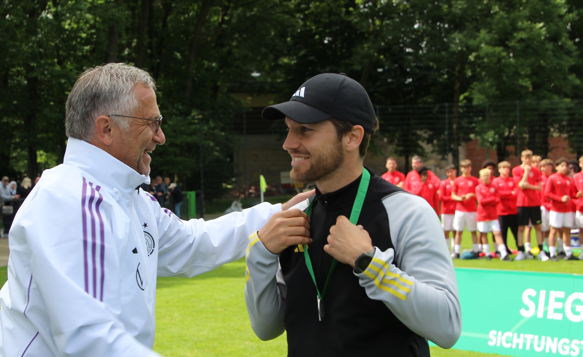 [{"clang_id":1,"value":"Zwei Männer, vermutlich Trainer und Spieler, begrüßen sich mit erhobenen Fäusten. Im Hintergrund steht eine Gruppe in roten Trikots auf einem Fußballfeld."}]