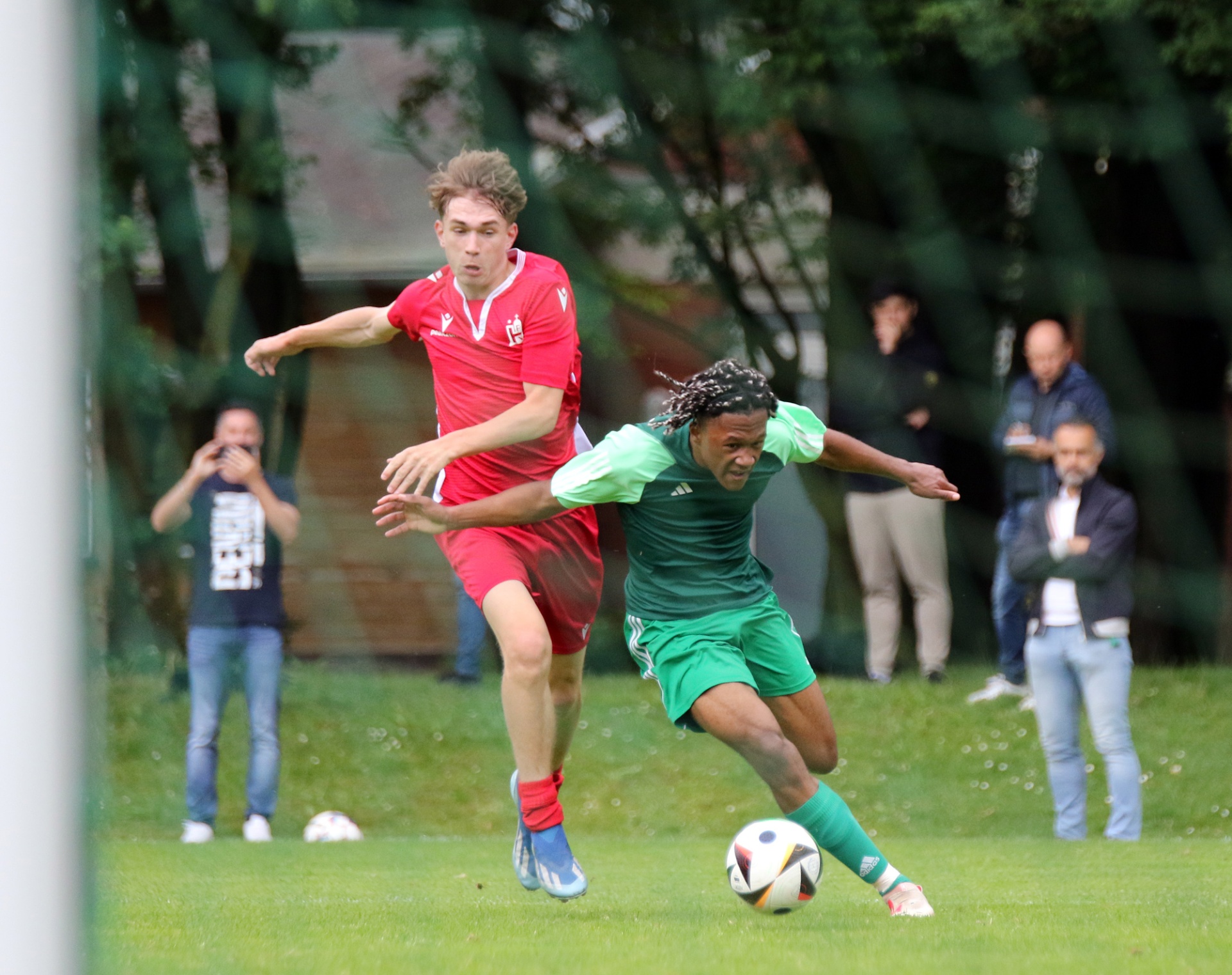 [{"clang_id":1,"value":"Zwei Fußballspieler im Wettkampf: Links in Rot, rechts in Grün, dribbeln den Ball auf einem grünen Rasenfeld. Zuschauer im Hintergrund."}]