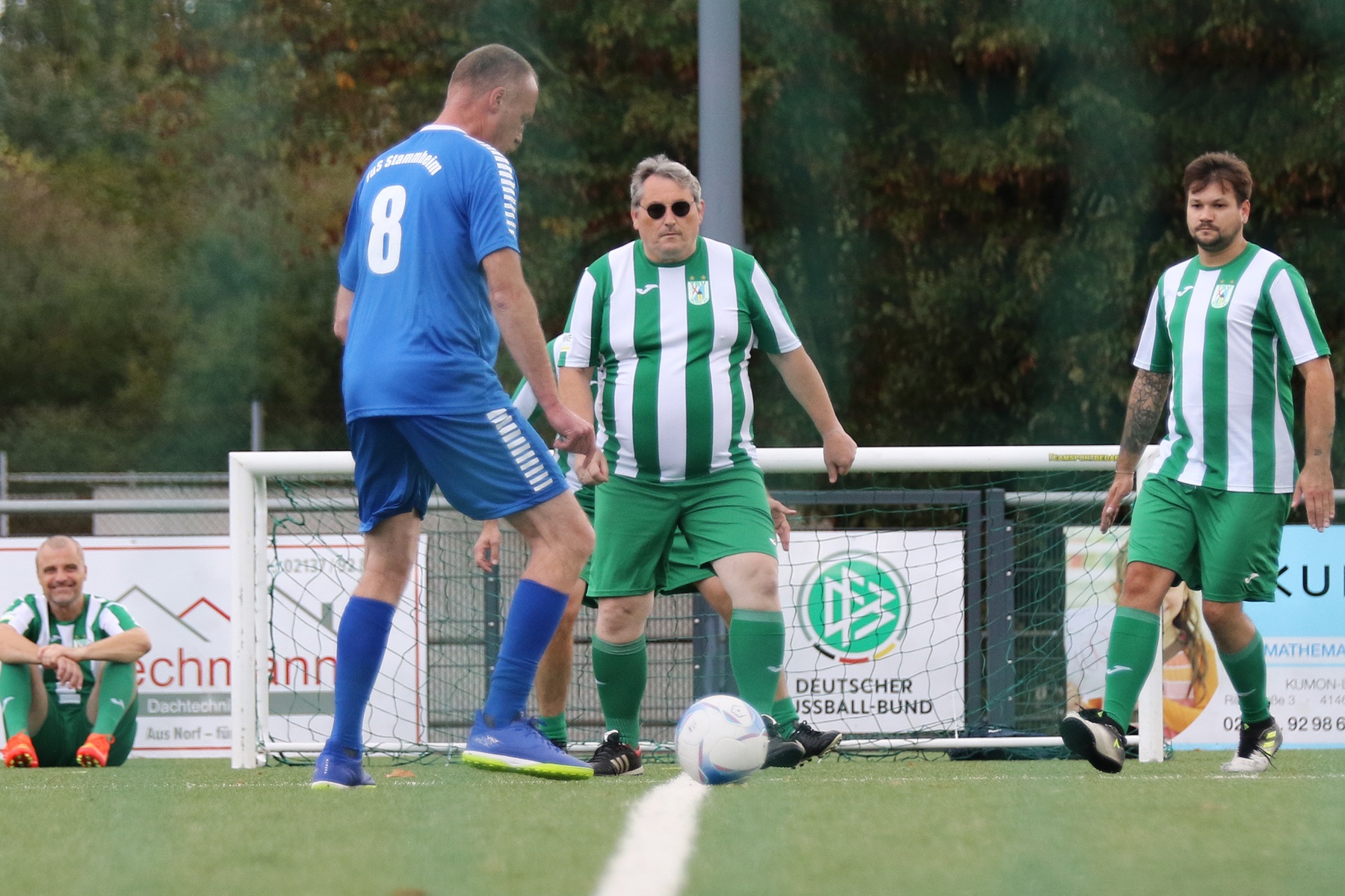 [{"clang_id":1,"value":"Fußballspiel: Spieler in Blau dribbelt den Ball an drei Gegnern in Grün-Weiß vorbei. Im Hintergrund eine Werbetafel und ein Zaun."}]