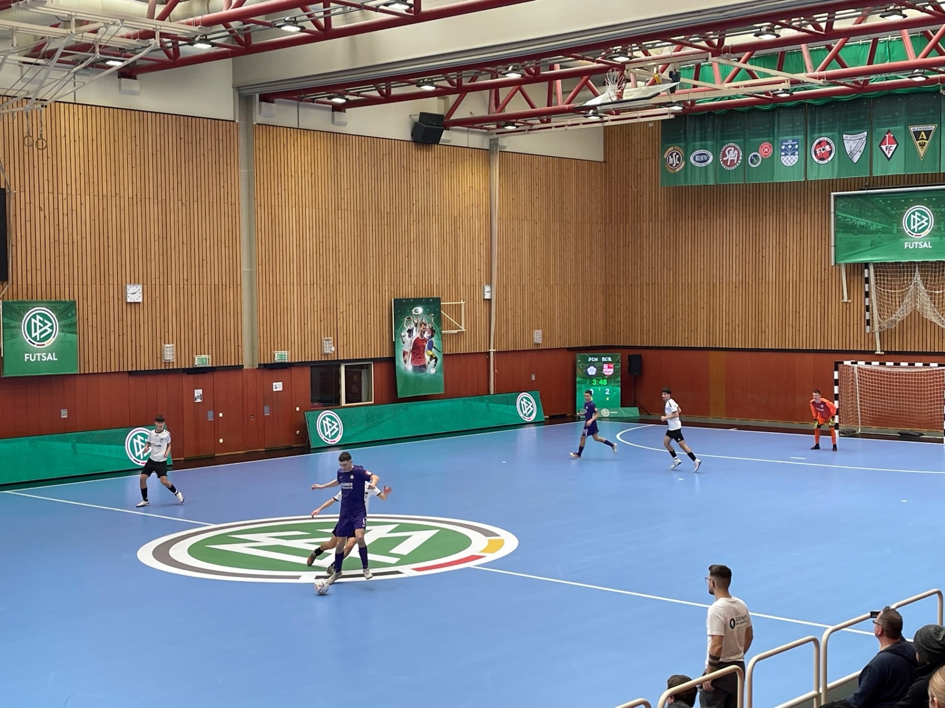 [{"clang_id":1,"value":"Futsal-Halle mit blauem Spielfeld und grünen Wandbereichen. Spieler in Aktion, Zuschauerreihen am rechten Bildrand. Banner und Logos an den Wänden."}]