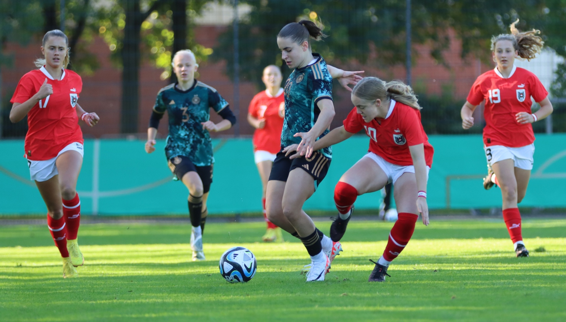 [{"clang_id":1,"value":"Fußballspiel. Mehrere Spielerinnen auf grünem Rasen, eine dribbelt den Ball. Rot- und dunkelgrün gemusterte Trikots. Im Hintergrund Zuschauer und Bäume."}]