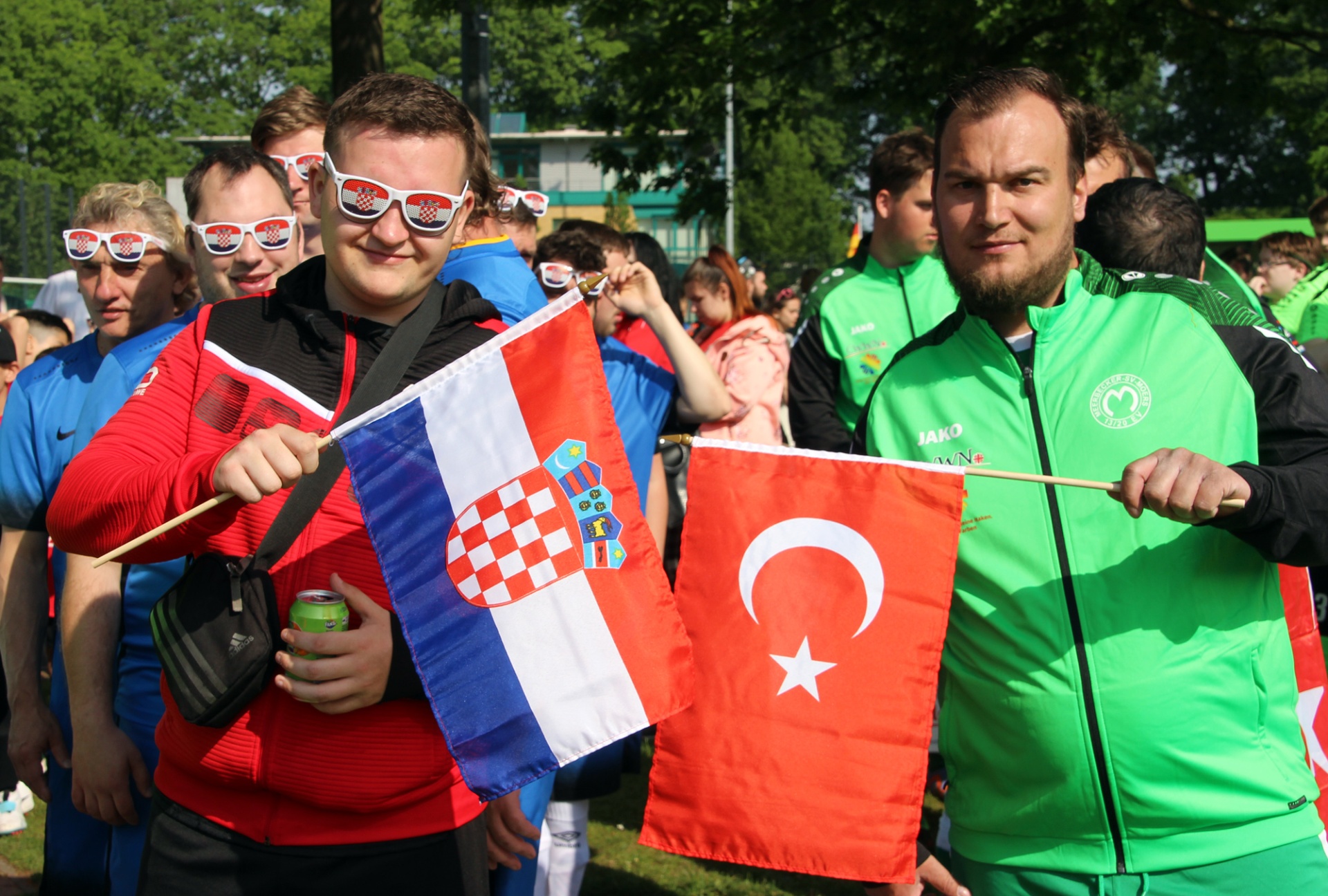 [{"clang_id":1,"value":"Zwei Männer halten Fahnen: links die kroatische, rechts die türkische. Im Hintergrund Menschen in Sportkleidung und mit Sonnenbrillen."}]