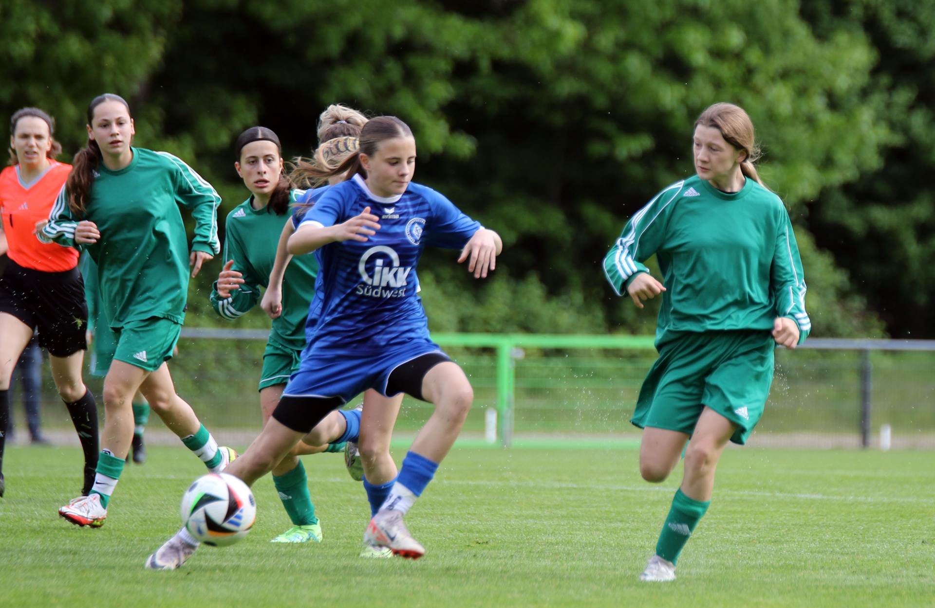 [{"clang_id":1,"value":"Fußballspiel: Jugendspielerinnen dribbeln und laufen auf einem grünen Rasenplatz. Spielerinnen tragen blaue und grüne Trikots. Bäume im Hintergrund."}]