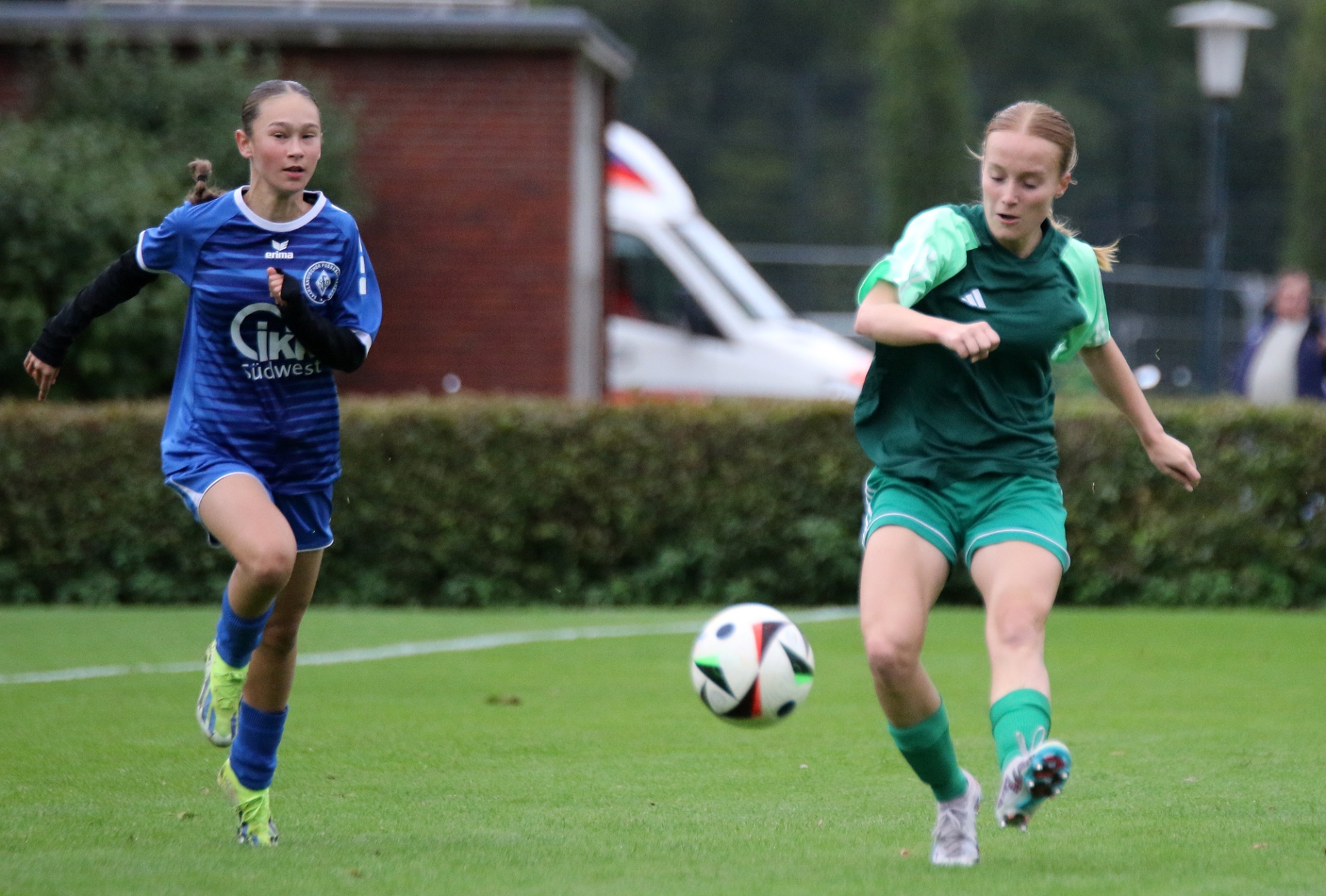 [{"clang_id":1,"value":"Zwei junge Fußballspielerinnen auf Grün. Links in blauem Trikot, rechts in grünem Trikot, beide in Bewegung und dem Ball zugewandt. Im Hintergrund Gebäude und Bäume."}]