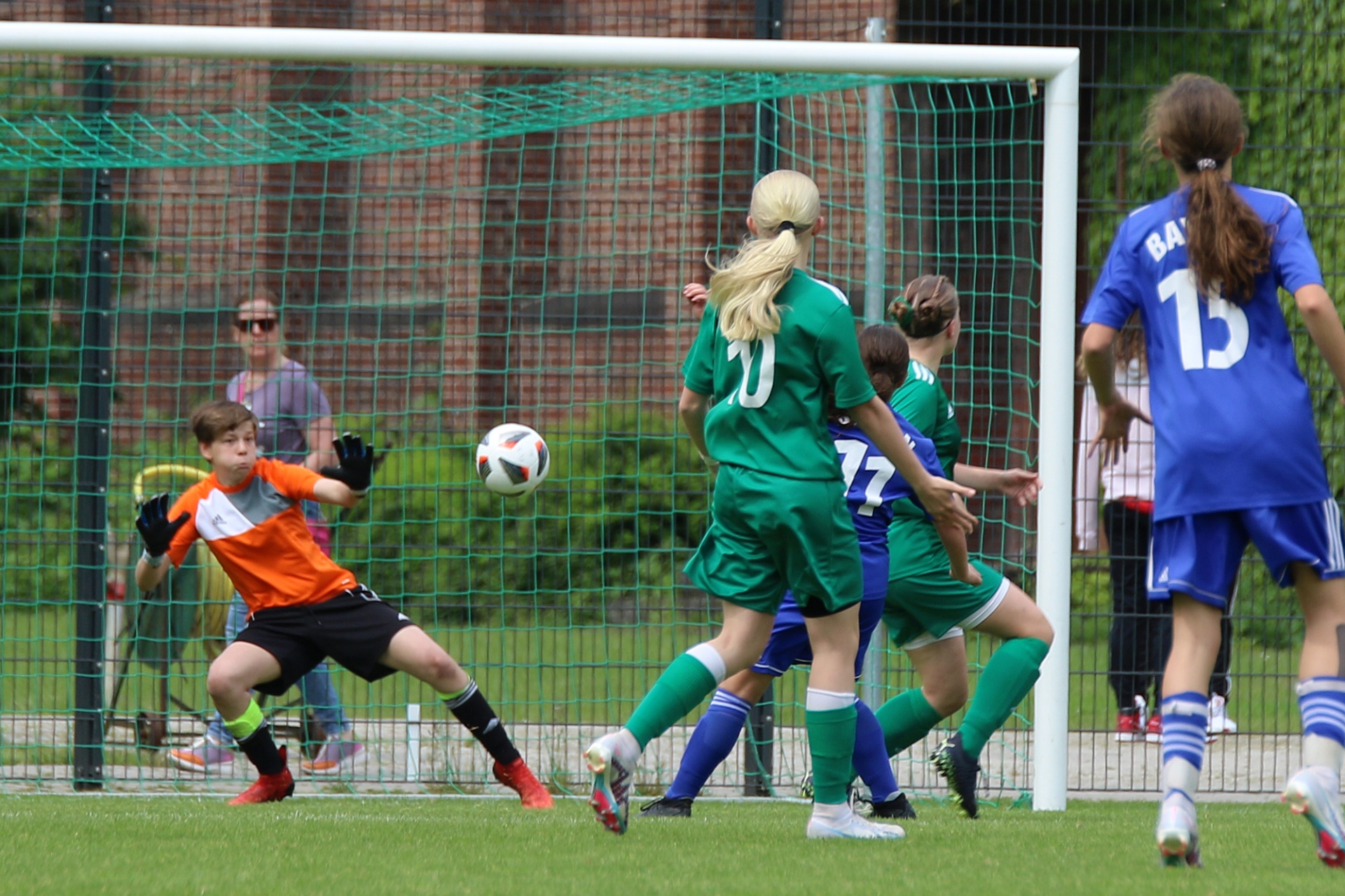 [{"clang_id":1,"value":"Fußballspiel: Torwart in orange wehrt Ball ab, mehrere Spielerinnen in grün und blau kämpfen um den Ball vor dem Tor. Brick Mauer im Hintergrund."}]