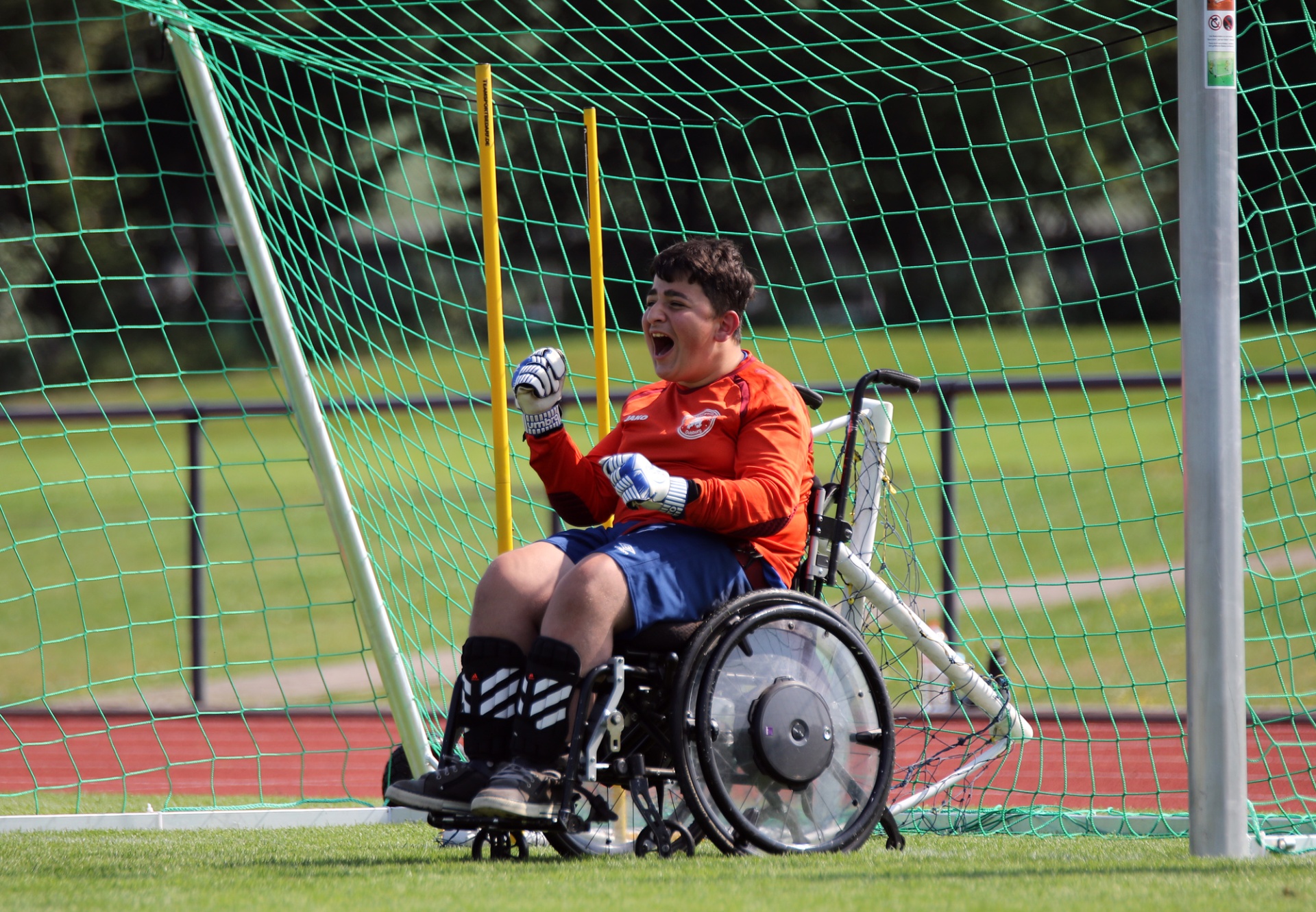 [{"clang_id":1,"value":"Rollstuhl-Fußballtorwart in rotem Trikot und blauen Shorts jubelt in seinem Sportrollstuhl vor dem Tornetz. Rasen und Laufbahn im Hintergrund."}]