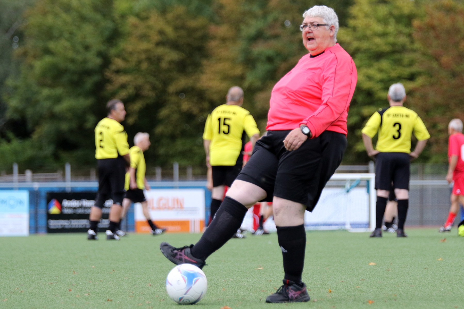 [{"clang_id":1,"value":"Ältere Frau mit Brille und rotem Oberteil schießt auf einem Fußballfeld einen Ball. Im Hintergrund weitere Spieler mit Trikots."}]