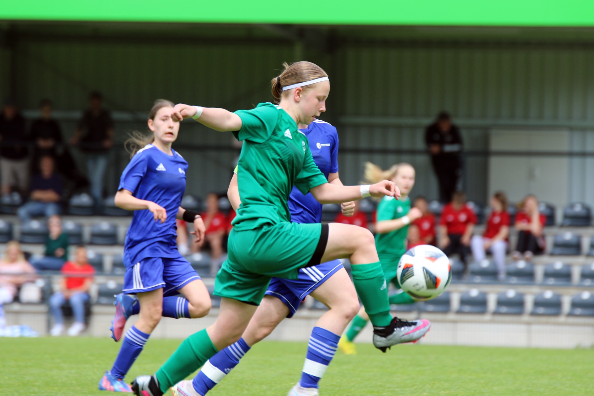 [{"clang_id":1,"value":"Zwei Fußballspielerinnen im Wettkampf. Eine in grünem Trikot schießt auf den Ball, eine andere in blauem Trikot rennt im Hintergrund. Zuschauer auf Tribünen."}]