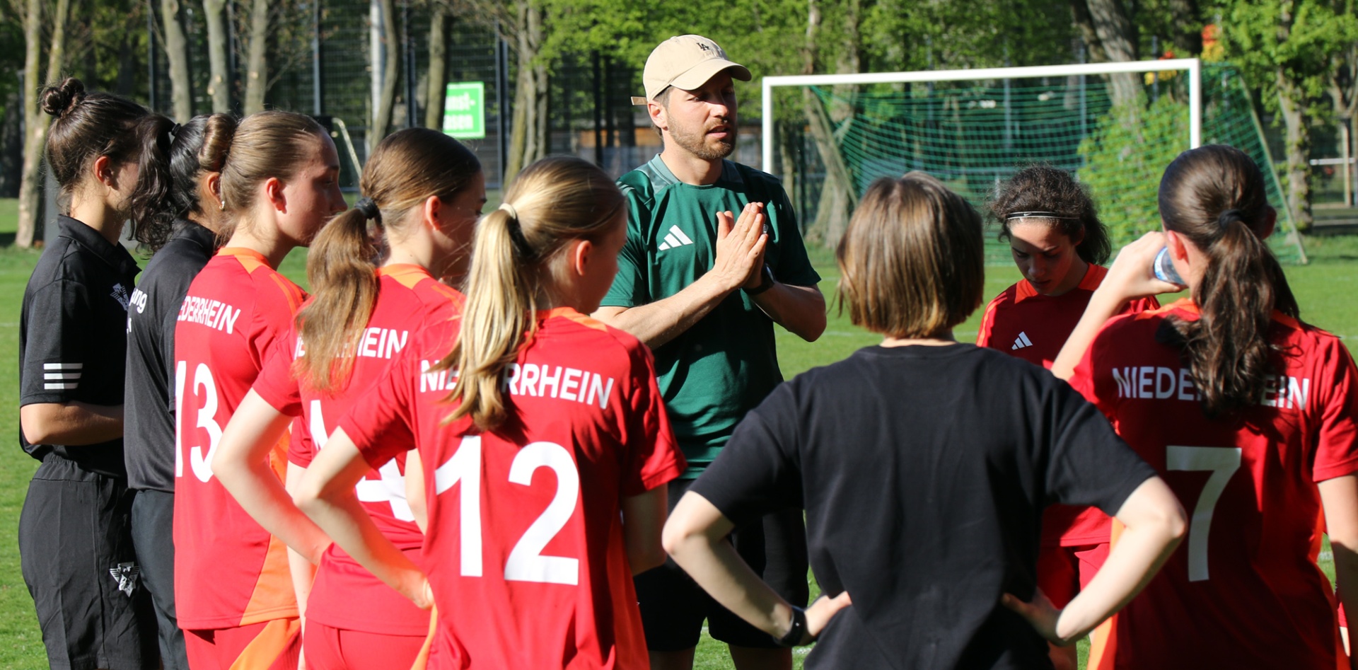 [{"clang_id":1,"value":"Fußballteam im Kreis um Trainer: Frauen und Mädchen in roten Trikots und schwarzer Sportkleidung stehen auf einem Fußballplatz zusammen."}]