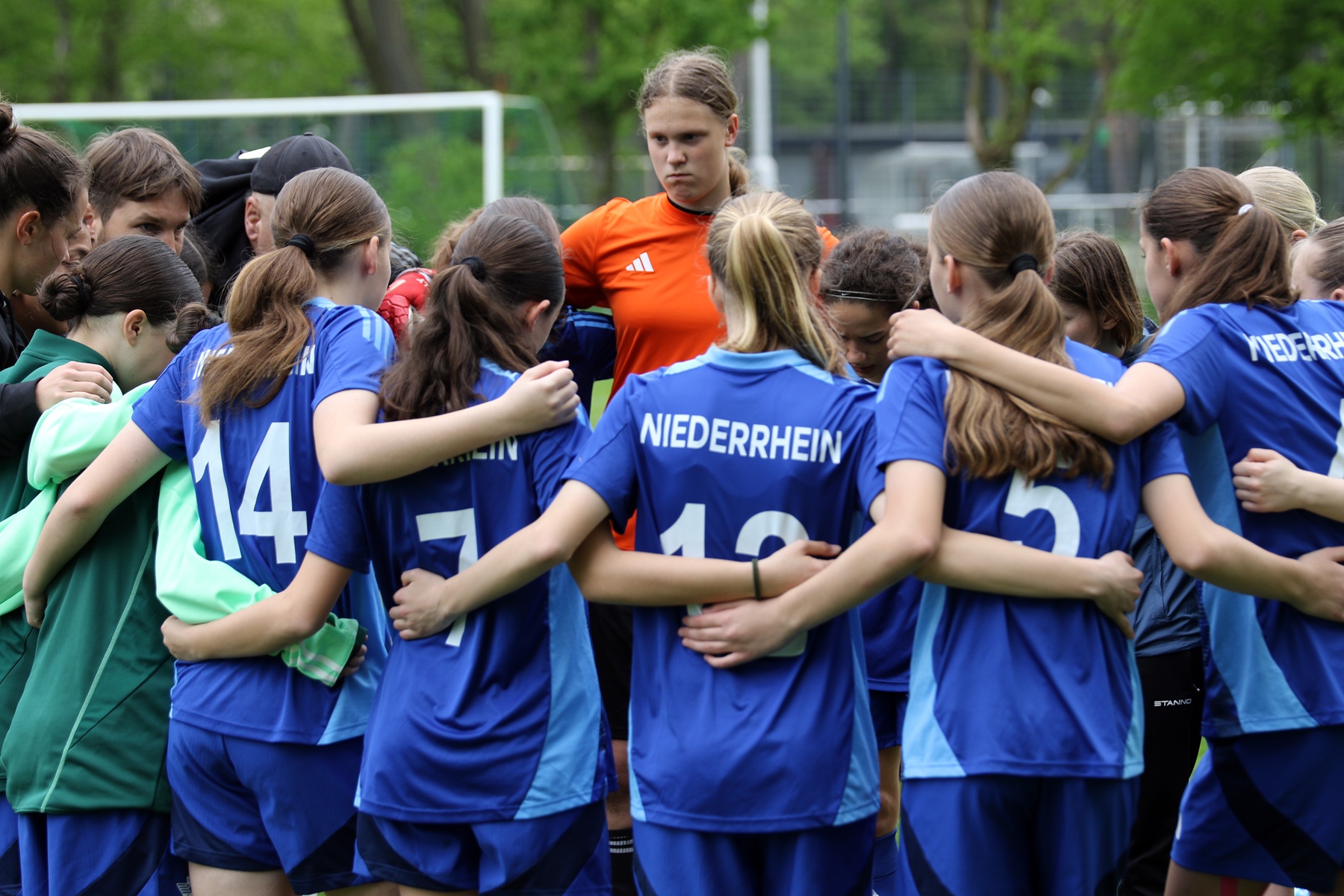 [{"clang_id":1,"value":"Fußballspielerinnen in blauen und grünen Trikots stehen im Kreis, Arme aneinander gelegt. Im Hintergrund eine Spielerin in orange und ein Fußballfeld."}]