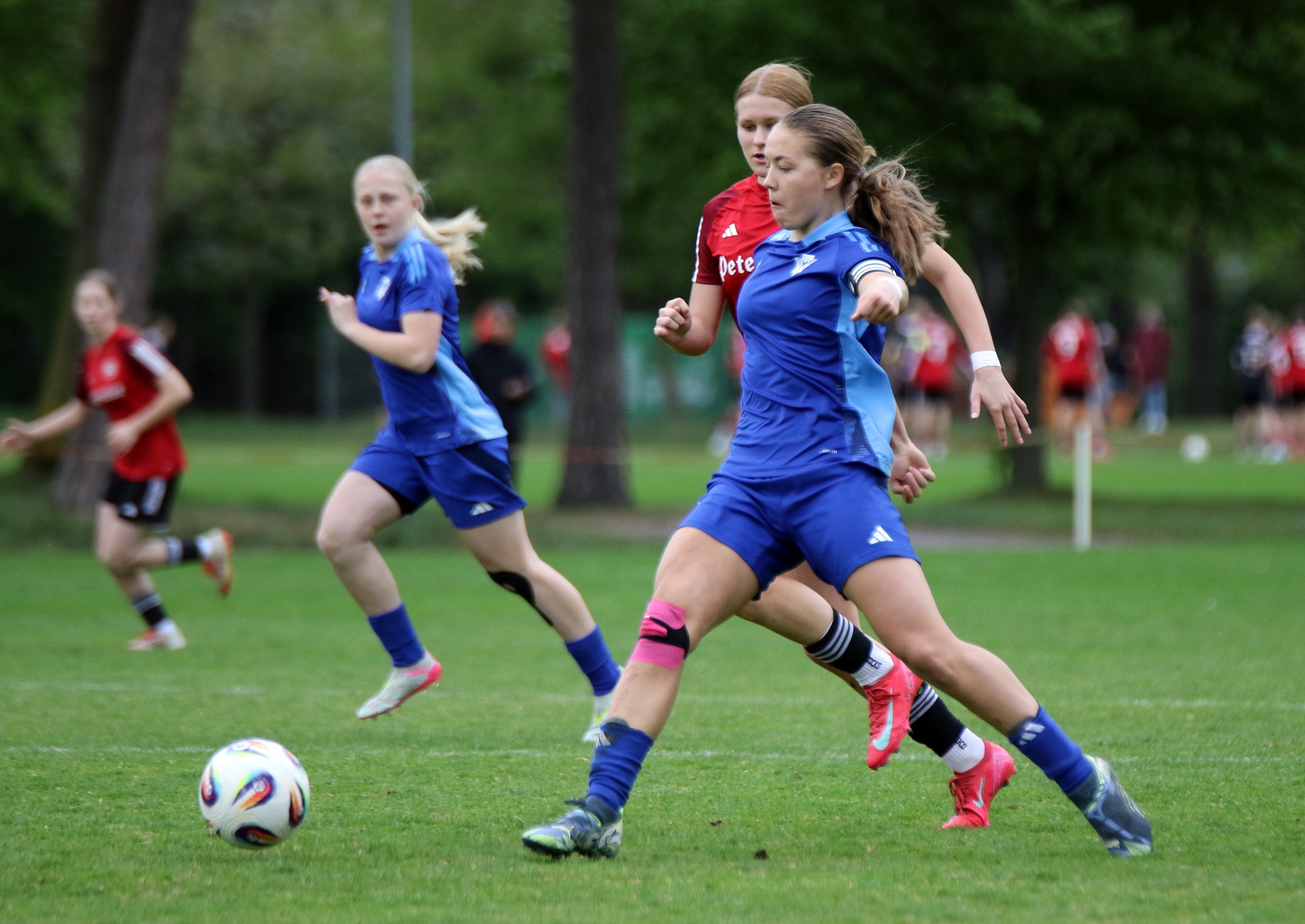 [{"clang_id":1,"value":"Fußballspiel: Drei Mädchen in blau und eine in rot laufen auf einem grünen Rasenfeld hinter einem Ball her. Bäume im Hintergrund."}]