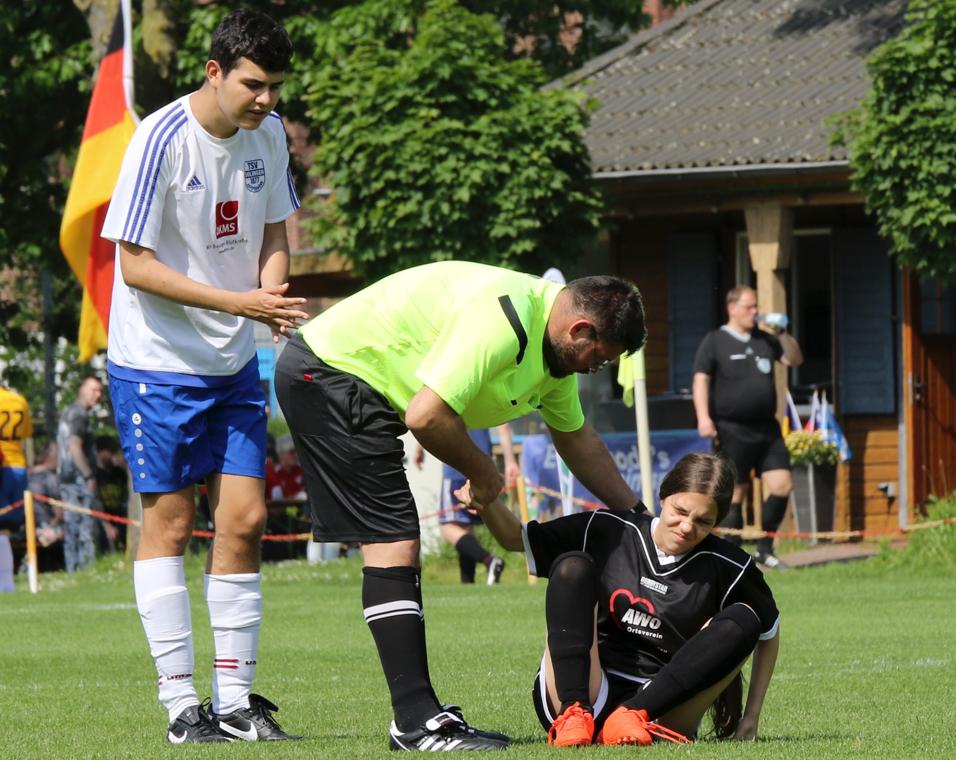 [{"clang_id":1,"value":"Fußballspiel: Spieler kniet am Boden, wird von Schiedsrichter und Mitspieler betreut. Im Hintergrund Zuschauer und ein Vereinsheim."}]