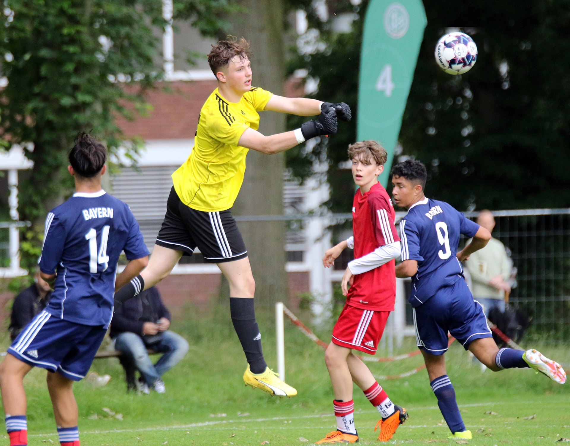 [{"clang_id":1,"value":"Fußballspiel: Torwart in Gelb springt hoch, um Ball abzufangen. Zwei Spieler in Blau und einer in Rot rennen in seine Richtung. Hintergrund: Zuschauer und grüne Bäume."}]