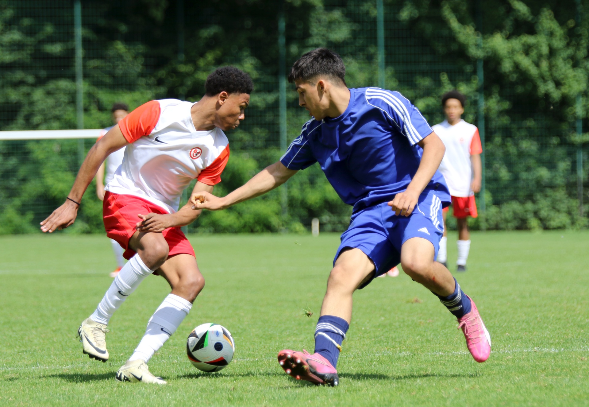 [{"clang_id":1,"value":"Zwei Jugendliche spielen Fußball auf einem grünen Rasen. Einer trägt rotes und weißes Trikot, der andere blaues. Ein Fußball liegt zwischen ihnen. Im Hintergrund weitere Personen und ein Zaun."}]