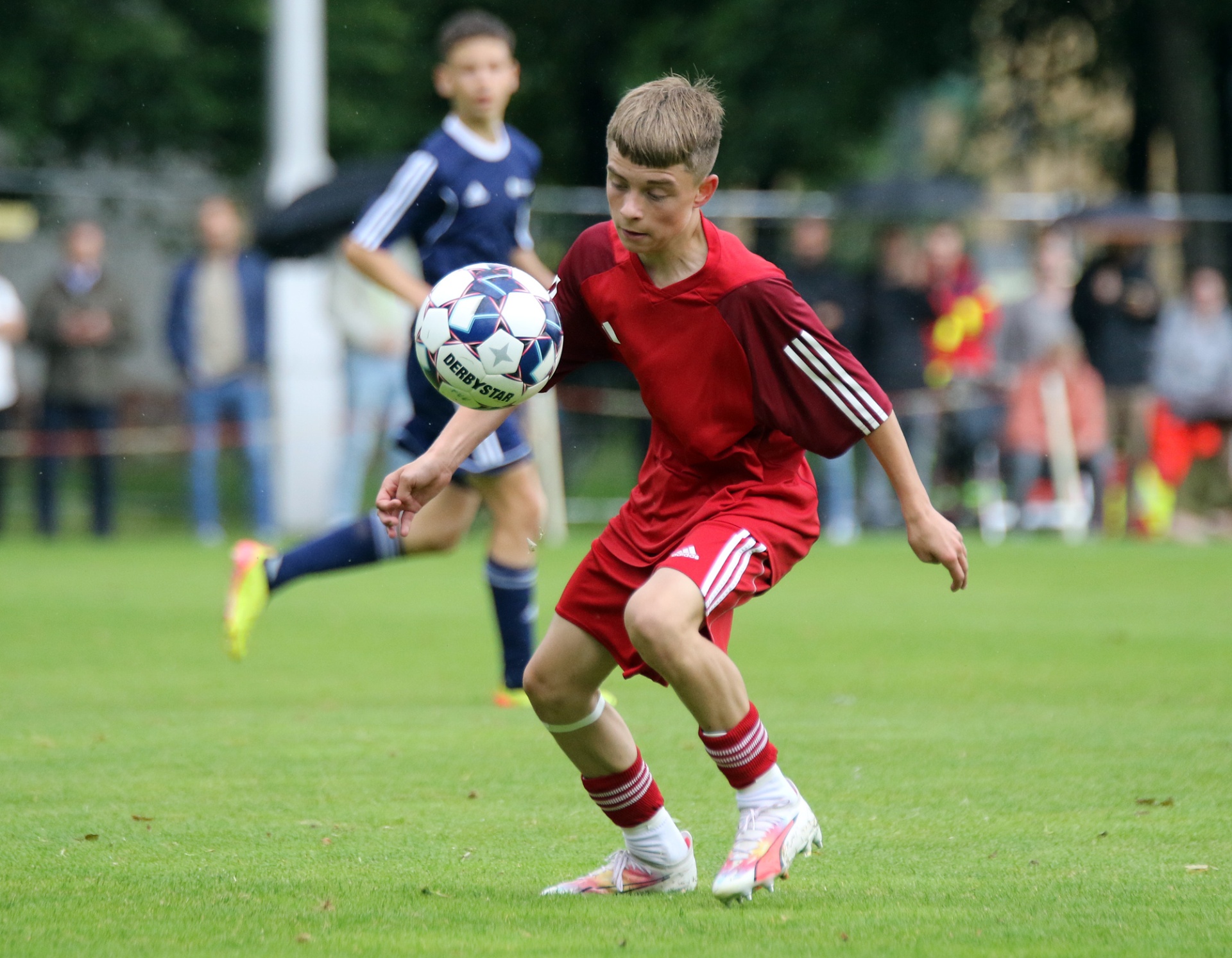 [{"clang_id":1,"value":"Zwei Fußballspieler auf grünem Rasen. Vorderer Spieler in rotem Trikot dribbelt den Ball, hinter ihm ein Spieler in blauem Trikot. Zuschauer im Hintergrund."}]
