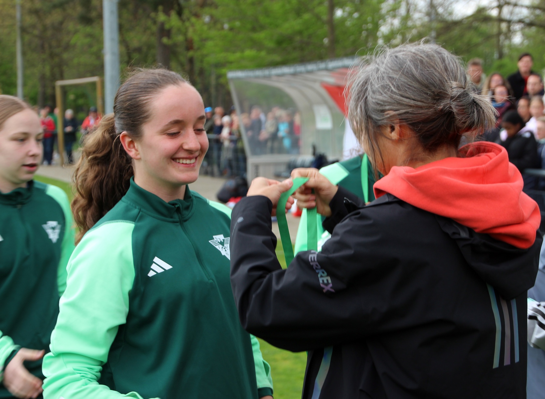 [{"clang_id":1,"value":"Zwei Frauen überreichen sich eine grüne Schleife auf einer Sportanlage. Im Hintergrund Zuschauer und ein Vereinsgebäude."}]
